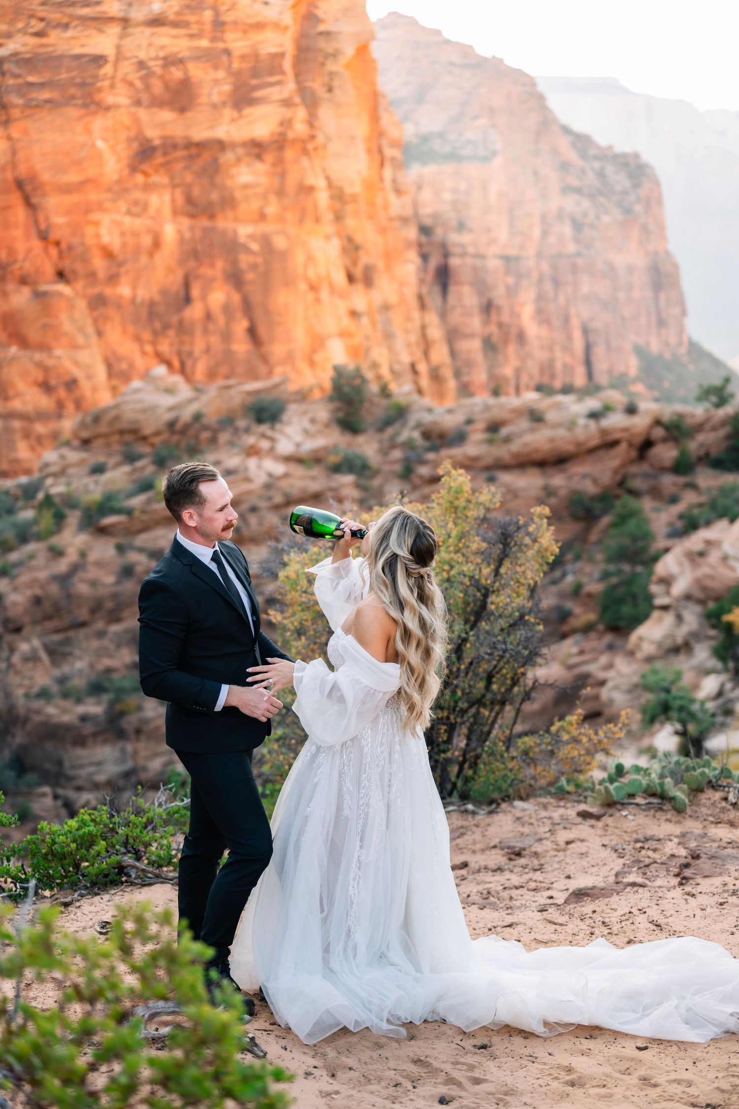 A couple celebrating with champagne against dramatic red canyon cliffs during sunset at outdoor wedding ceremony.