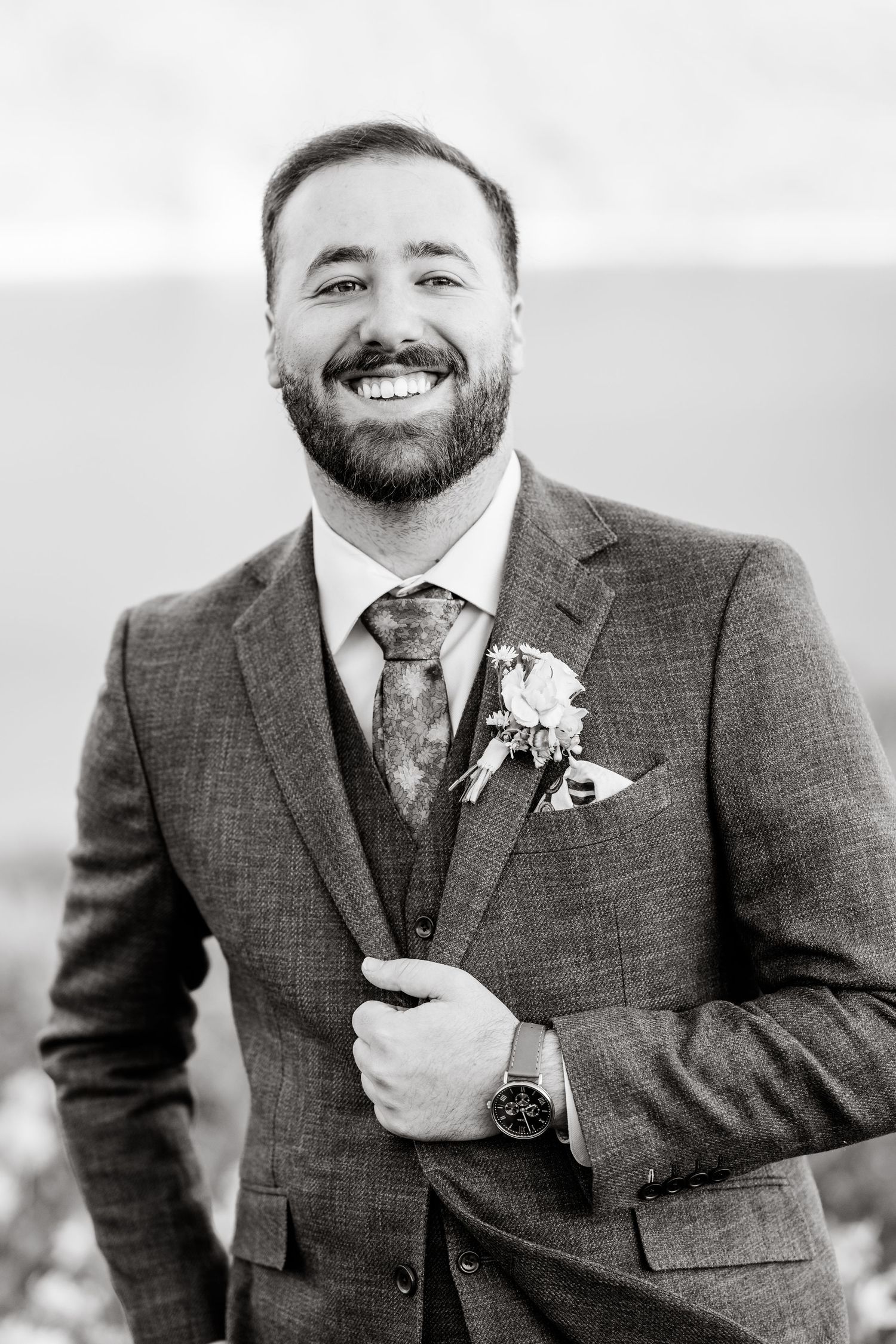 Portrait series showing groom in grey tweed suit with boutonniere against natural background.