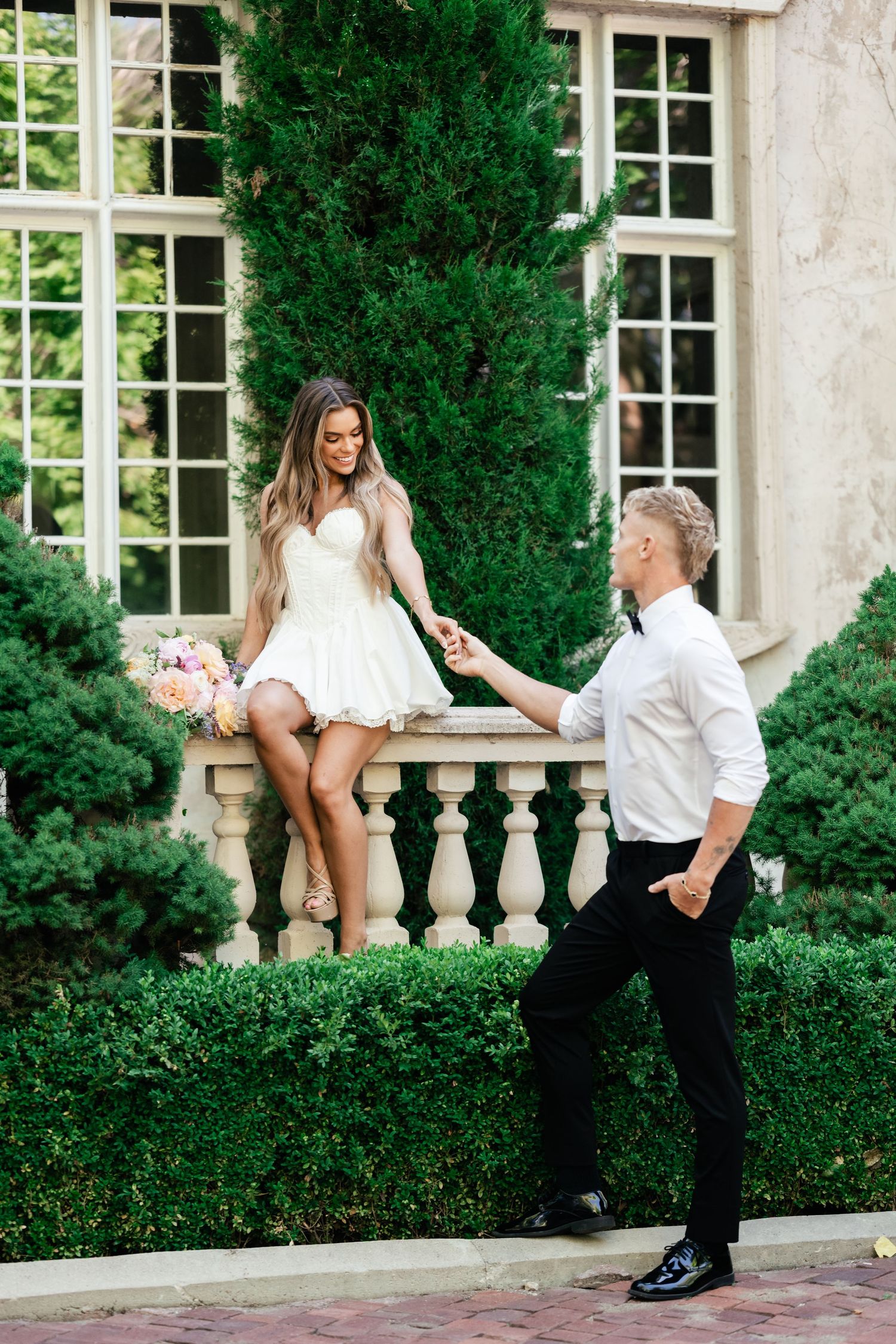 Romantic couple in formal attire pose together near stone balustrade with green cypress trees and French windows in background.