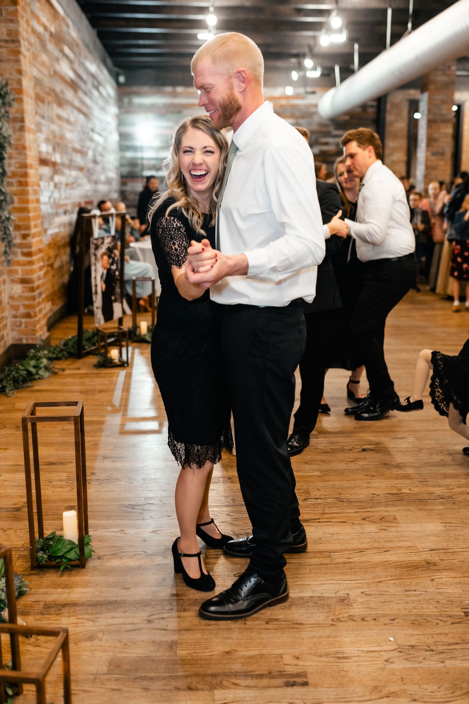 A couple dances joyfully on a wooden floor in an industrial-style venue with exposed brick walls and lantern decorations.