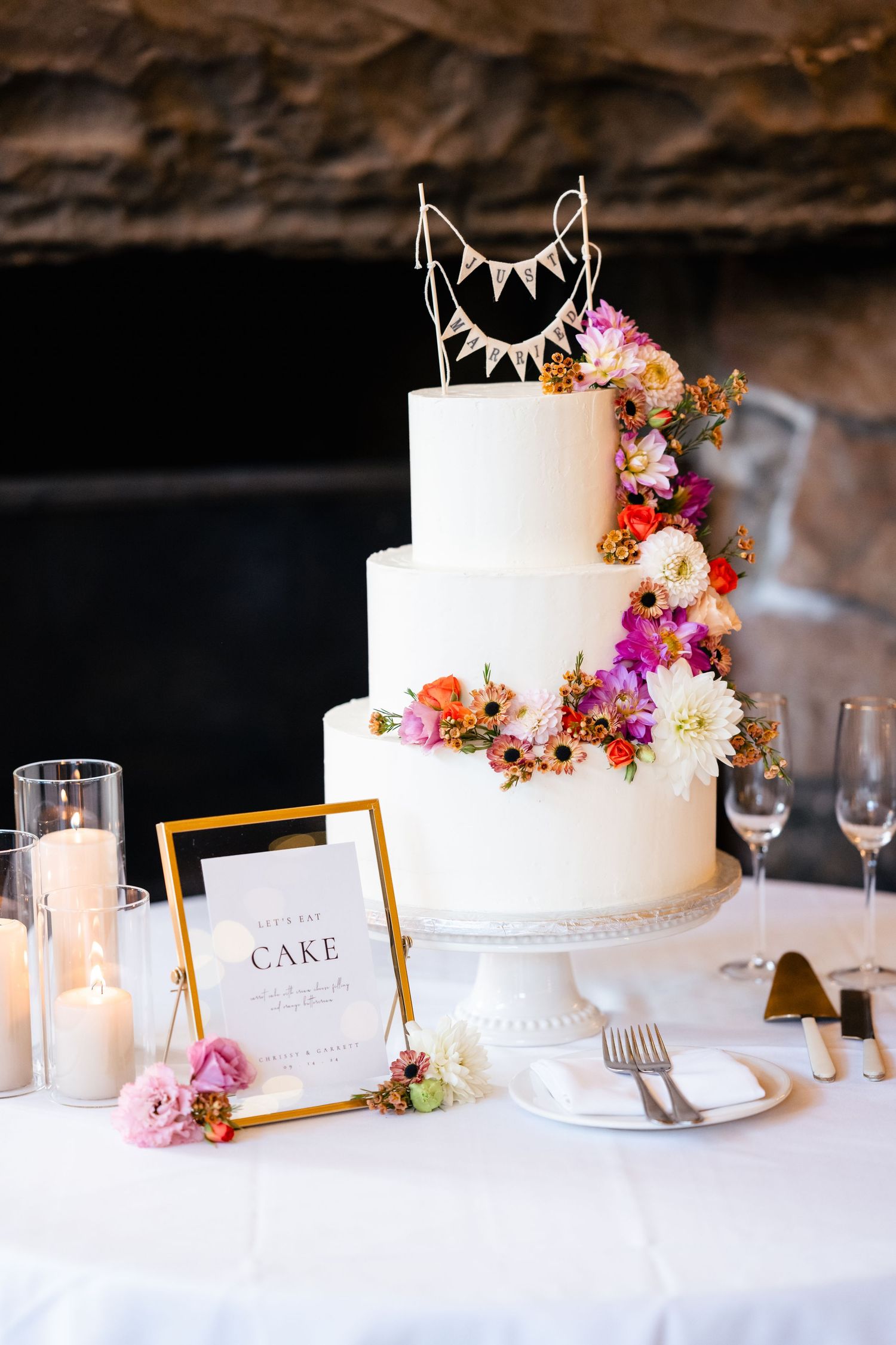 Elegant three-tier white wedding cake decorated with colorful fresh flowers and bunting topper on display with candles and menu card.