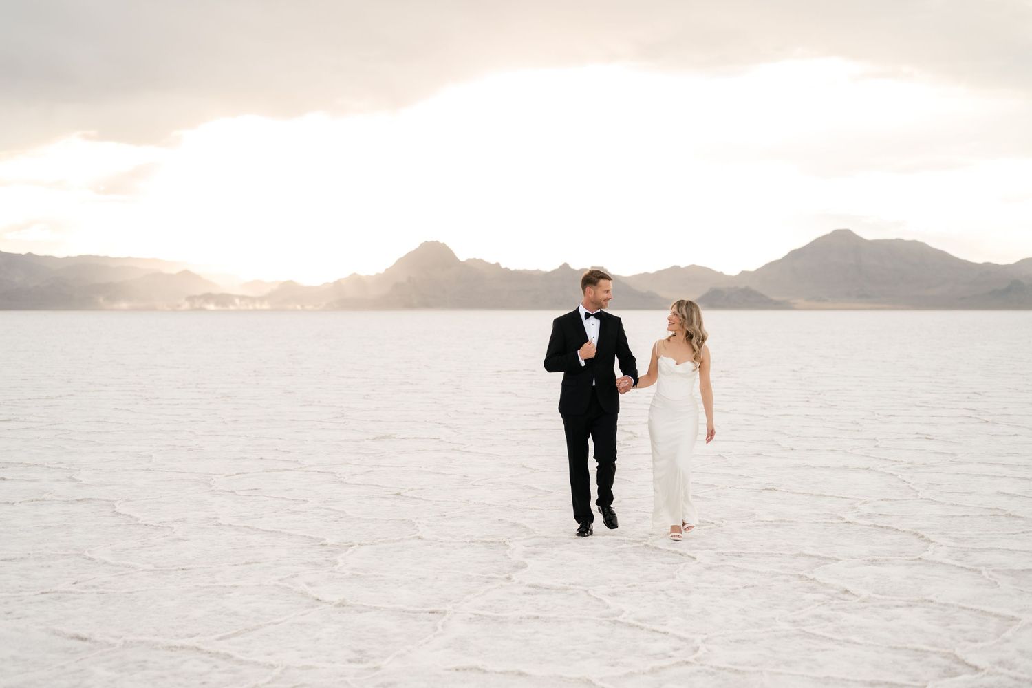A couple walks hand in hand across the stark white landscape of Bonneville Salt Flats at sunset. Salt Flats Wedding Photographer.