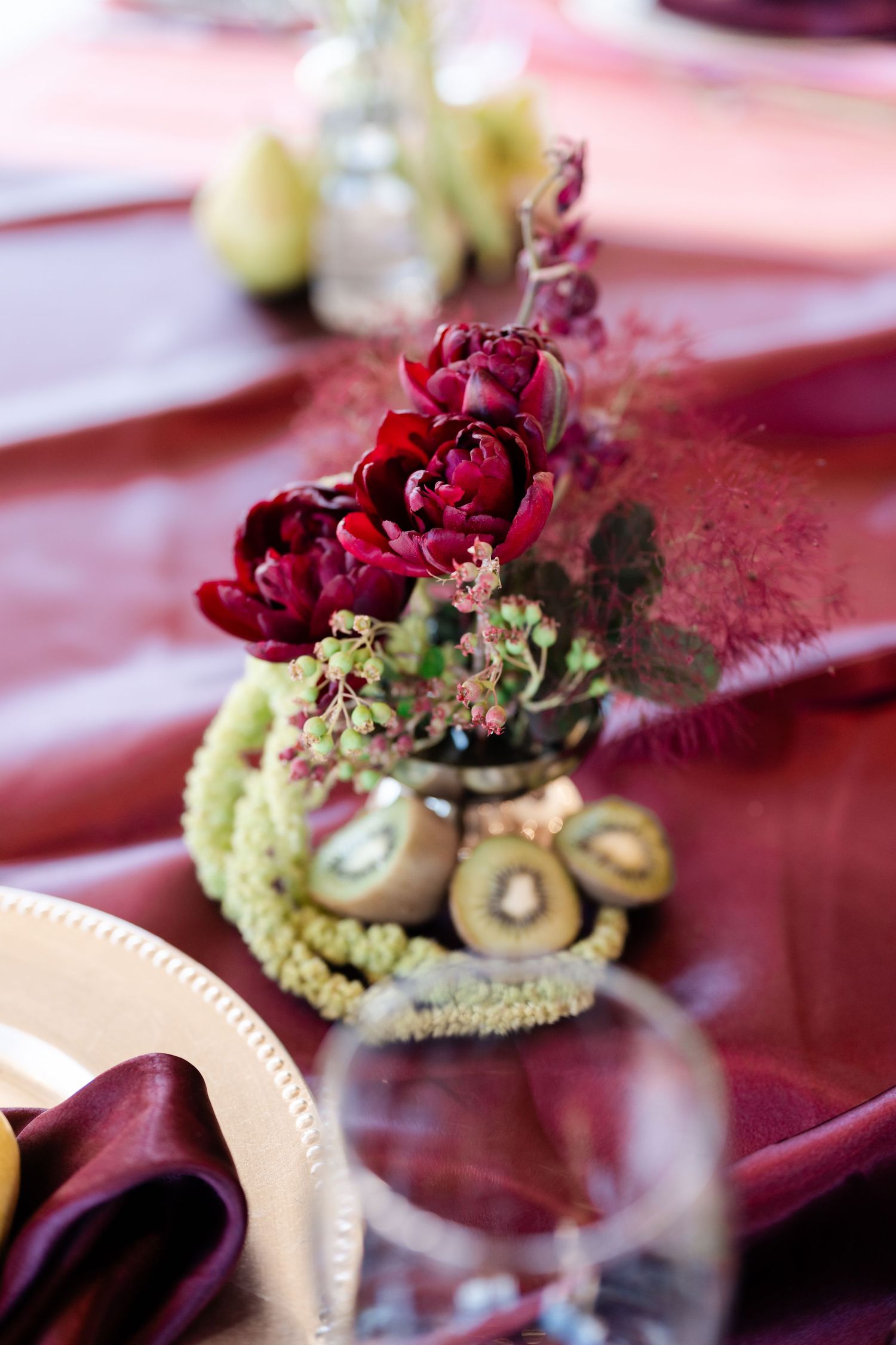 Elegant burgundy floral centerpiece with dark red peonies and kiwi fruit slices on a deep red satin tablecloth.