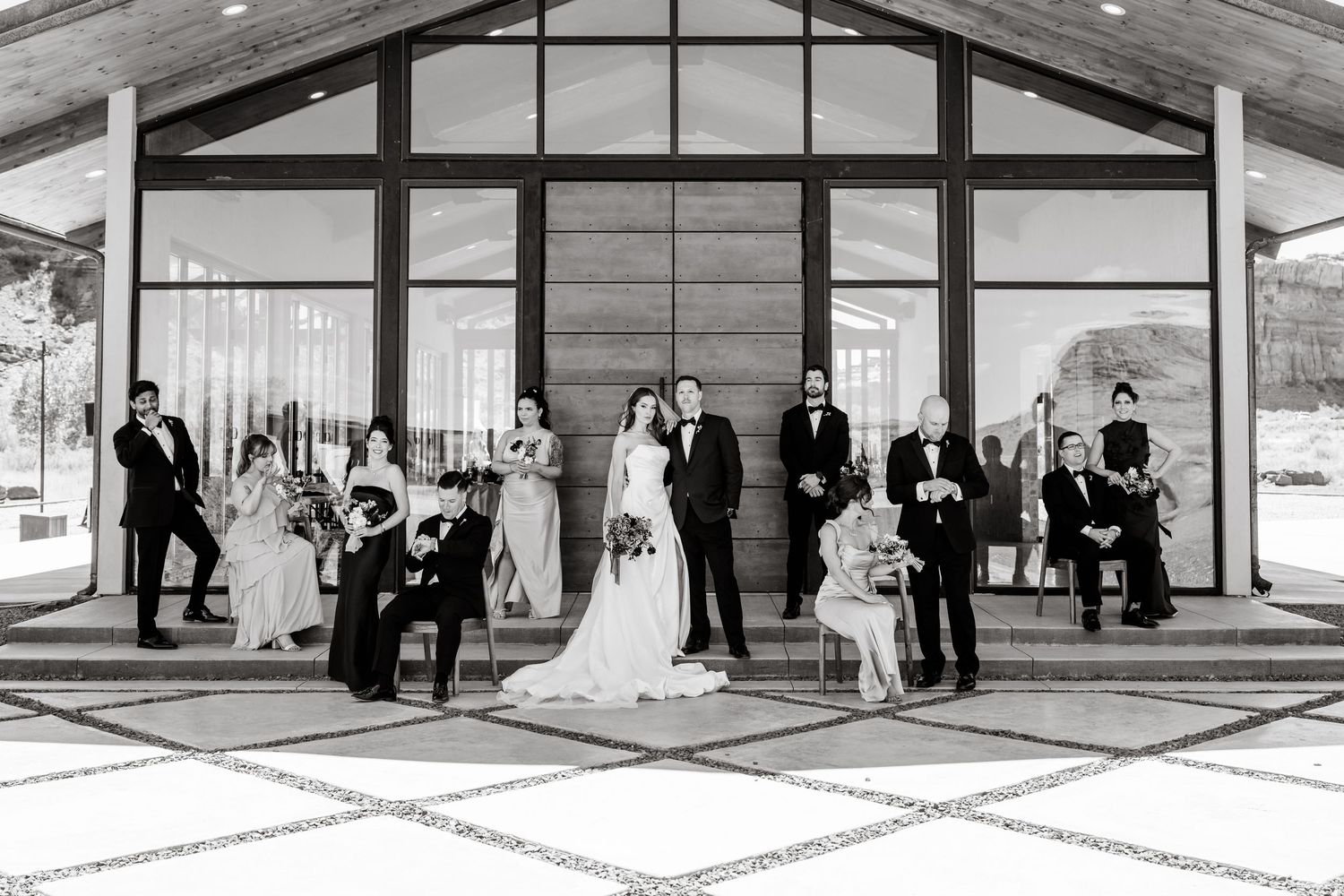 Black and white wedding party photo outside modern venue with bride, groom, and guests posing elegantly.