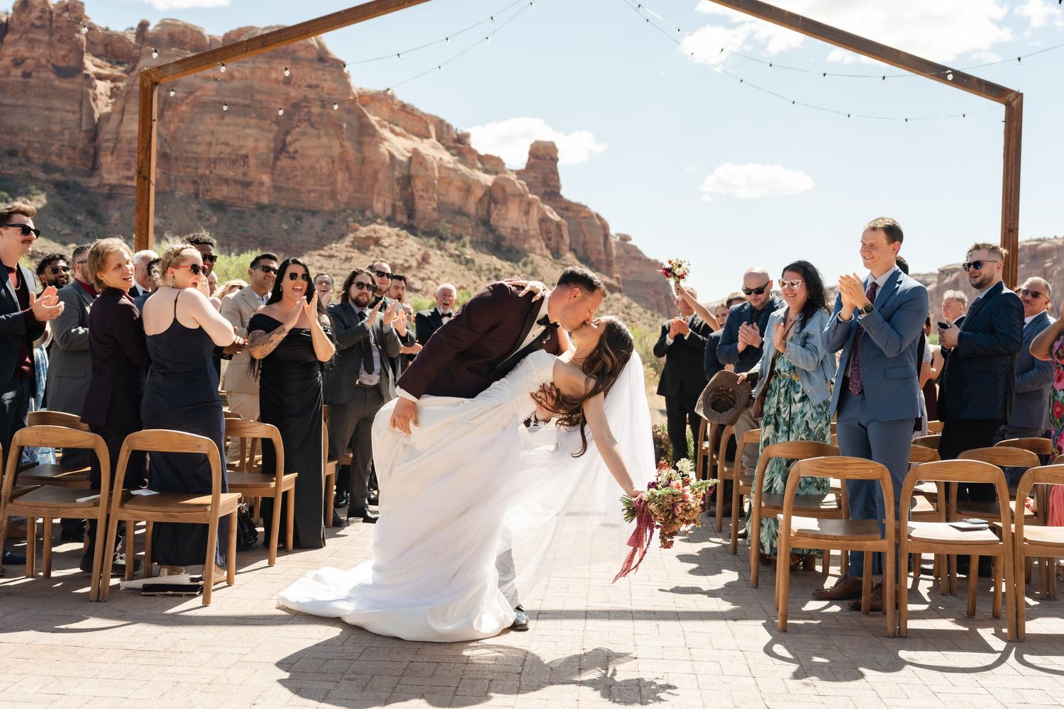 Couple shares romantic dip kiss at outdoor wedding ceremony with red rock canyon backdrop and cheering guests.