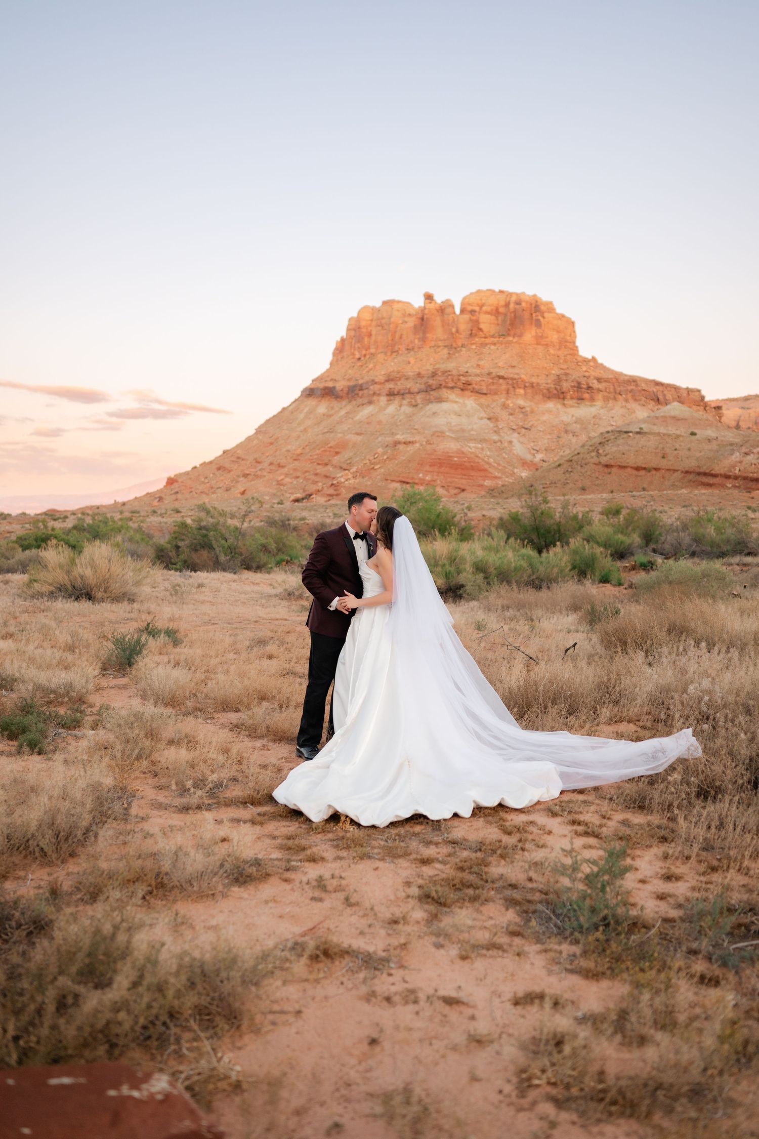 Bride and groom share a kiss in desert landscape with stunning red mesa butte glowing at sunset behind them.
