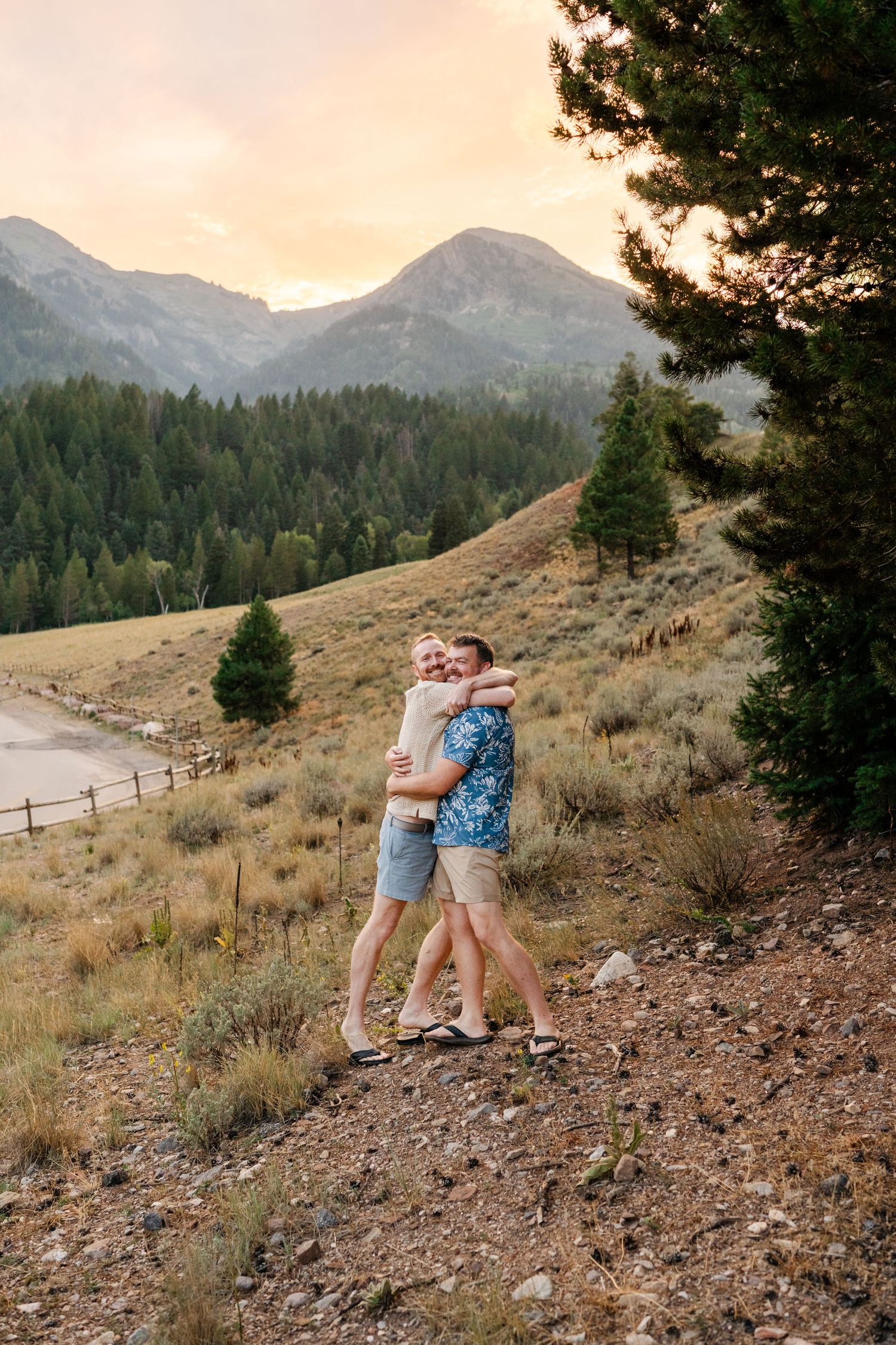 Couple hugging on a mountain hillside at sunset with pine trees and scenic peaks in the background.