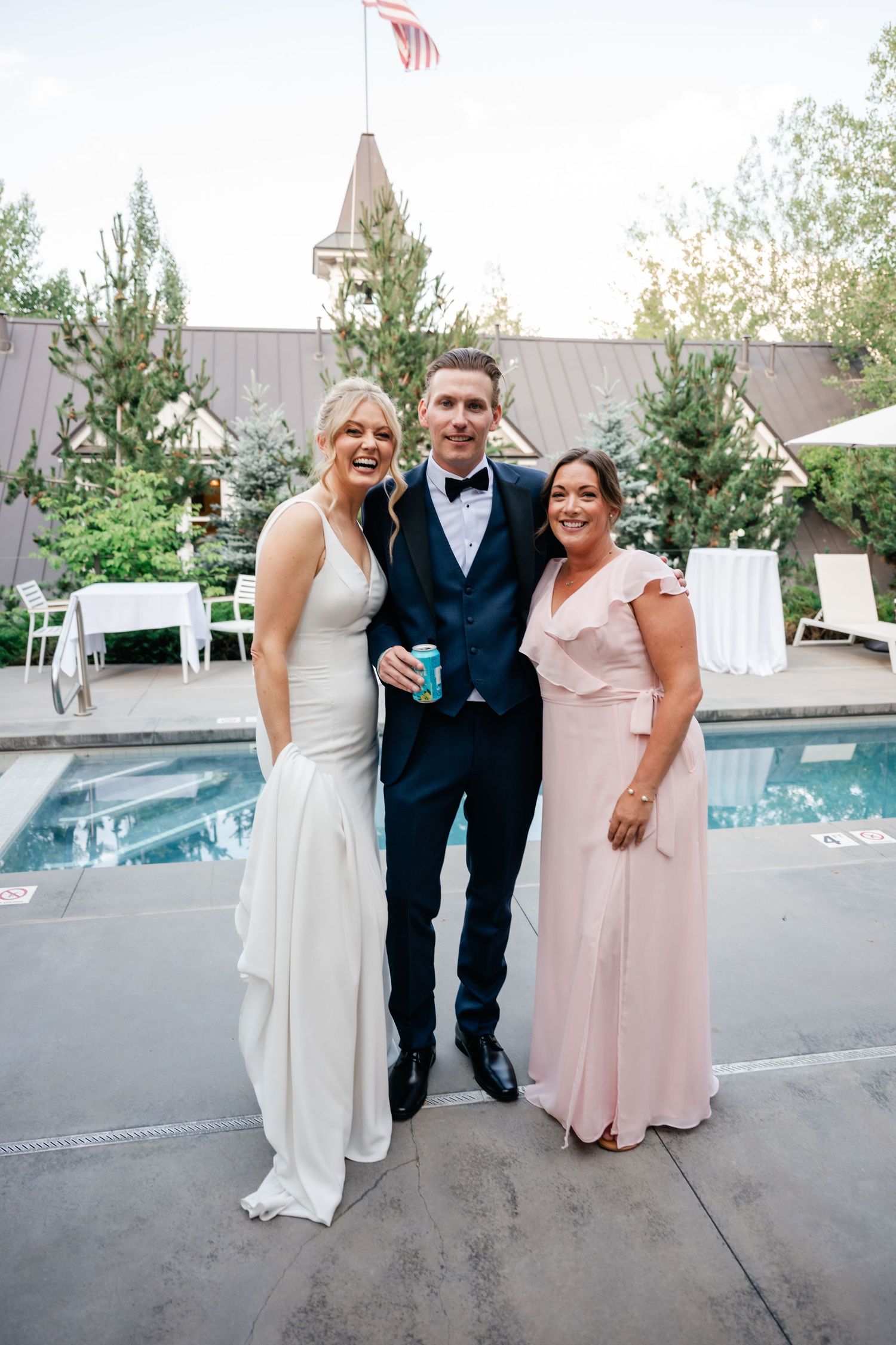 Bride in white gown and groom in navy tuxedo pose with guest in pink dress by outdoor pool at wedding reception.