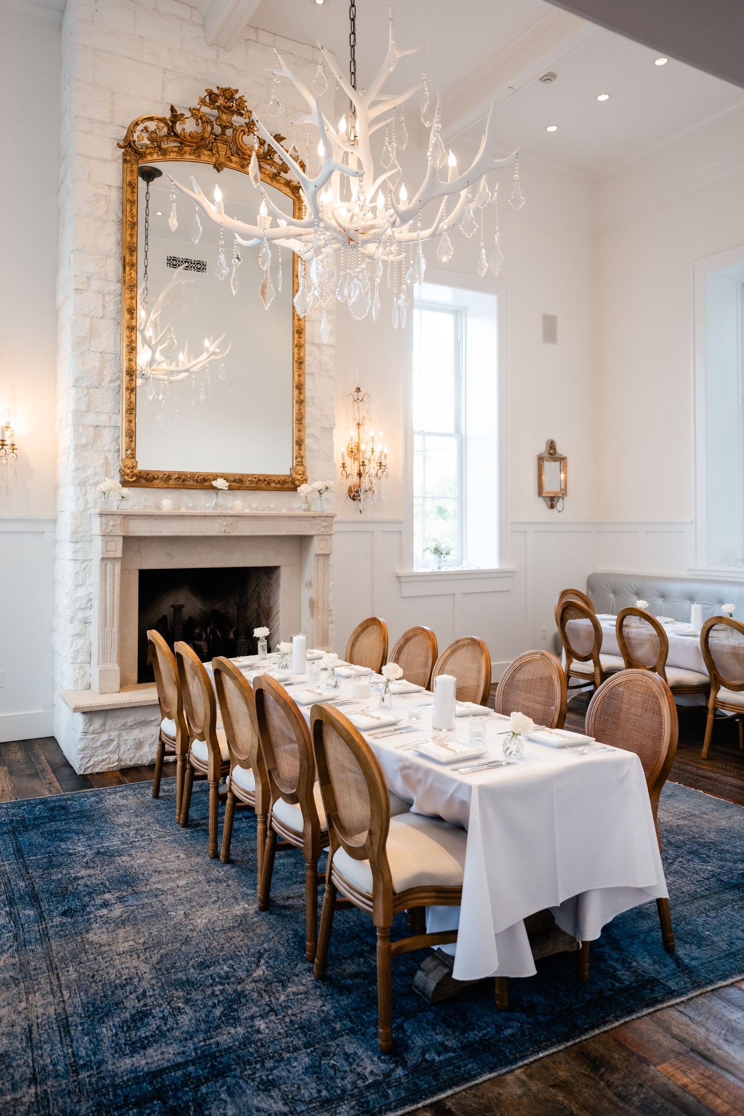 Elegant dining room with white antler chandelier, gold mirror, fireplace, long table with white linens, and French-style chairs on blue rug.