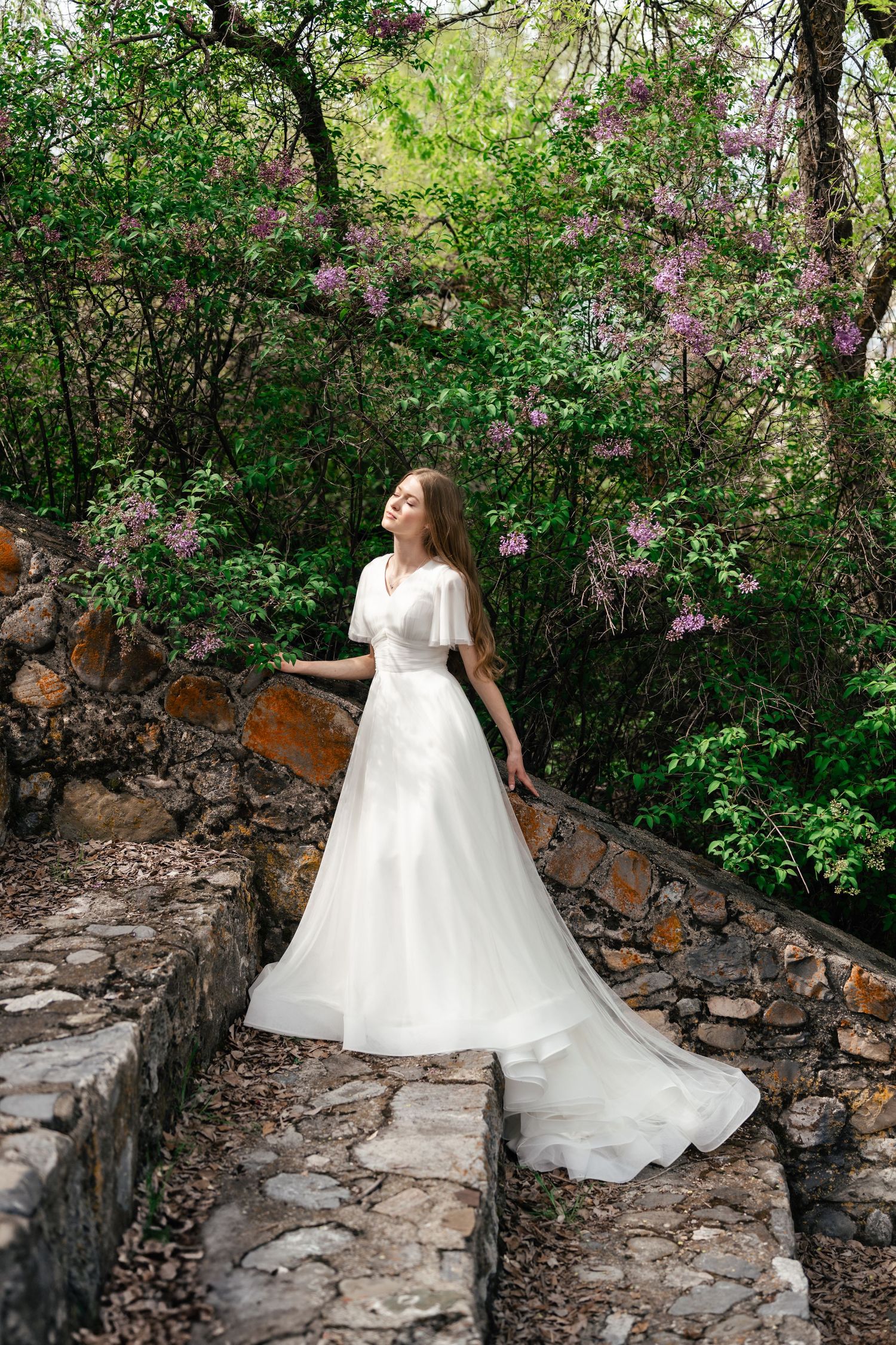 A bride in a flowing white wedding gown poses gracefully on stone steps surrounded by blooming lilac bushes in a garden setting.