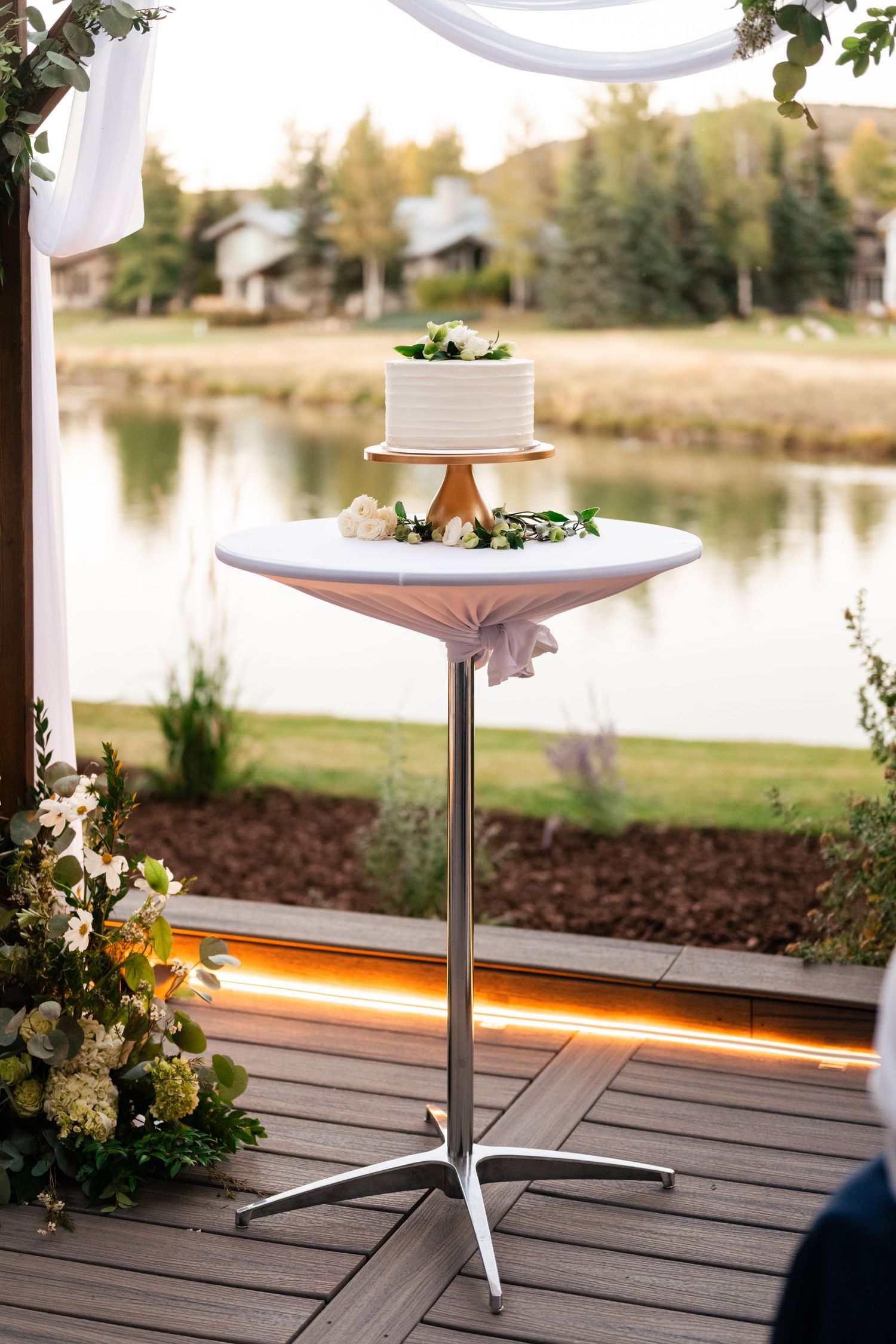 A white wedding cake display on a wooden deck overlooking a serene lake at sunset.