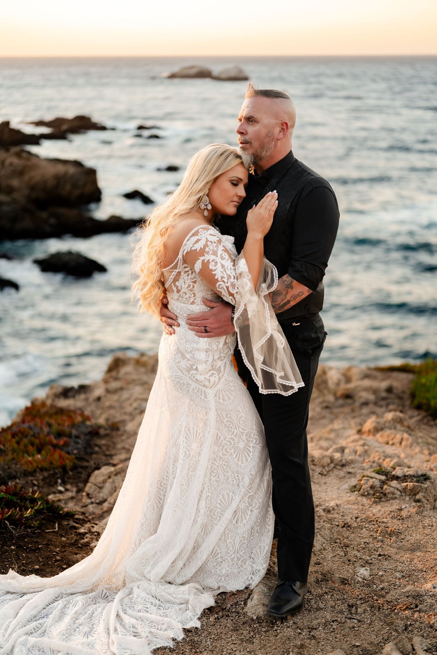 A couple in wedding attire dances intimately on a rocky coastal shoreline at sunset.