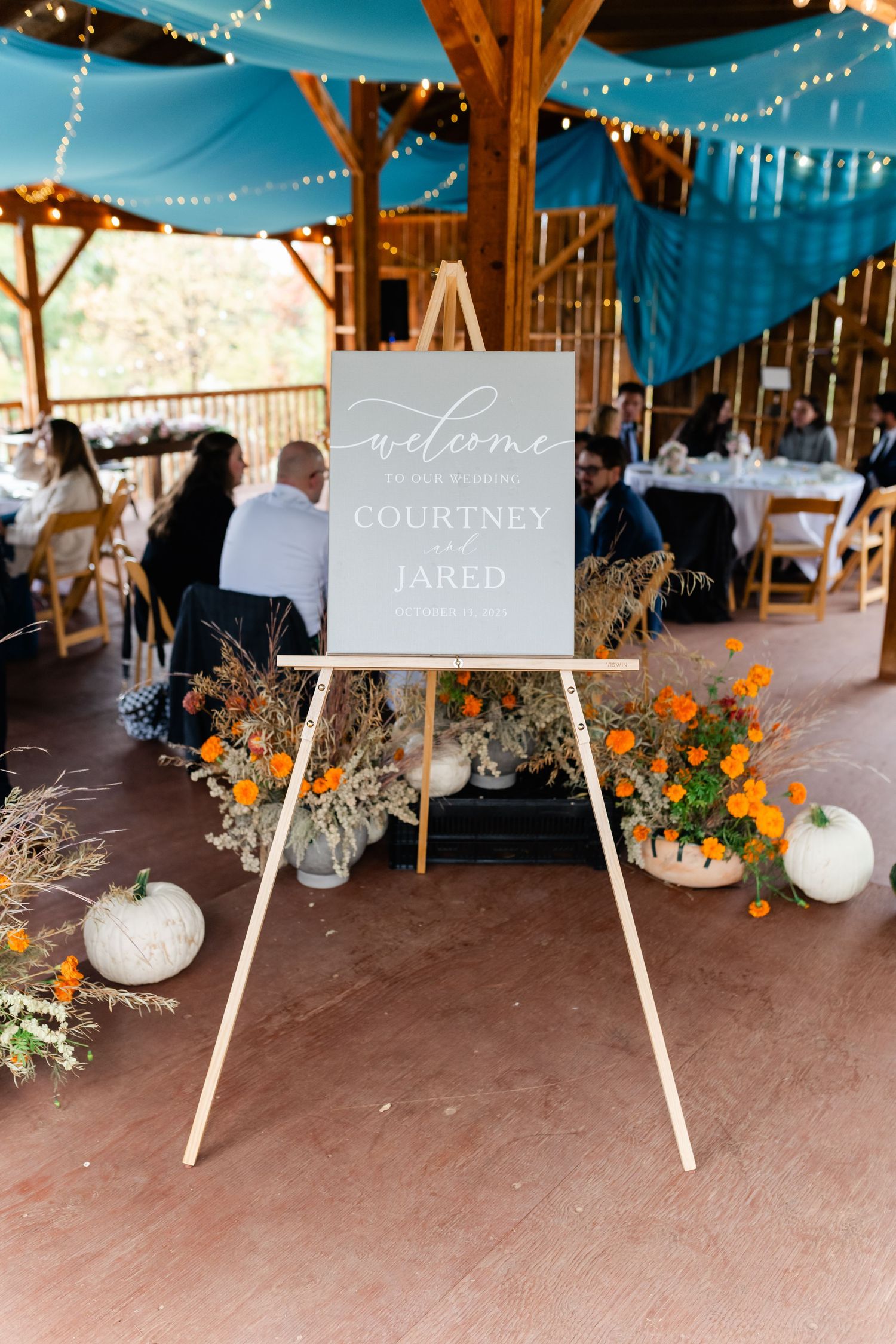 Rustic barn wedding reception setup with welcome signs and orange floral decorations under string lights.