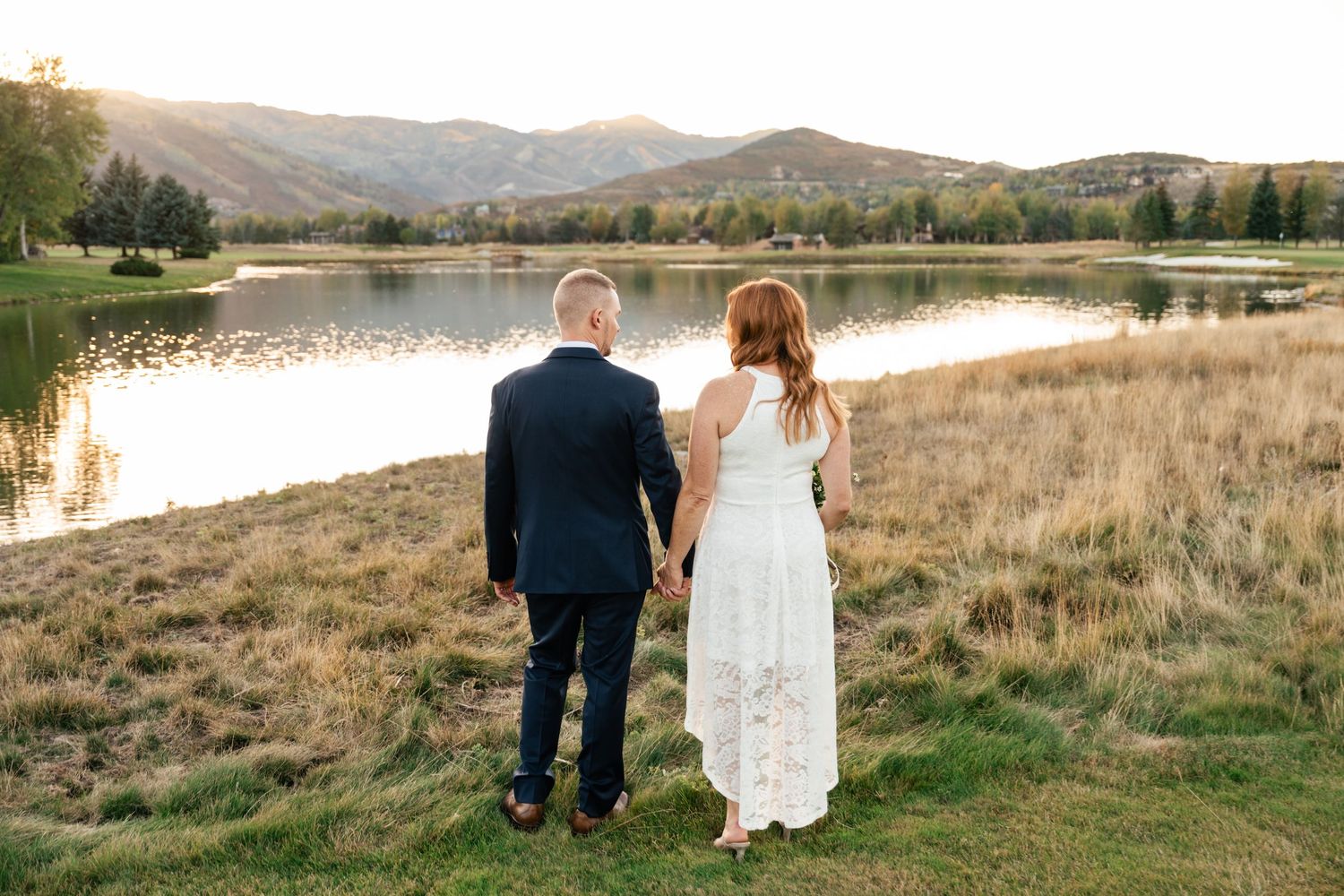 A couple walks hand in hand through a scenic mountain landscape with rolling hills and a lake in the background.