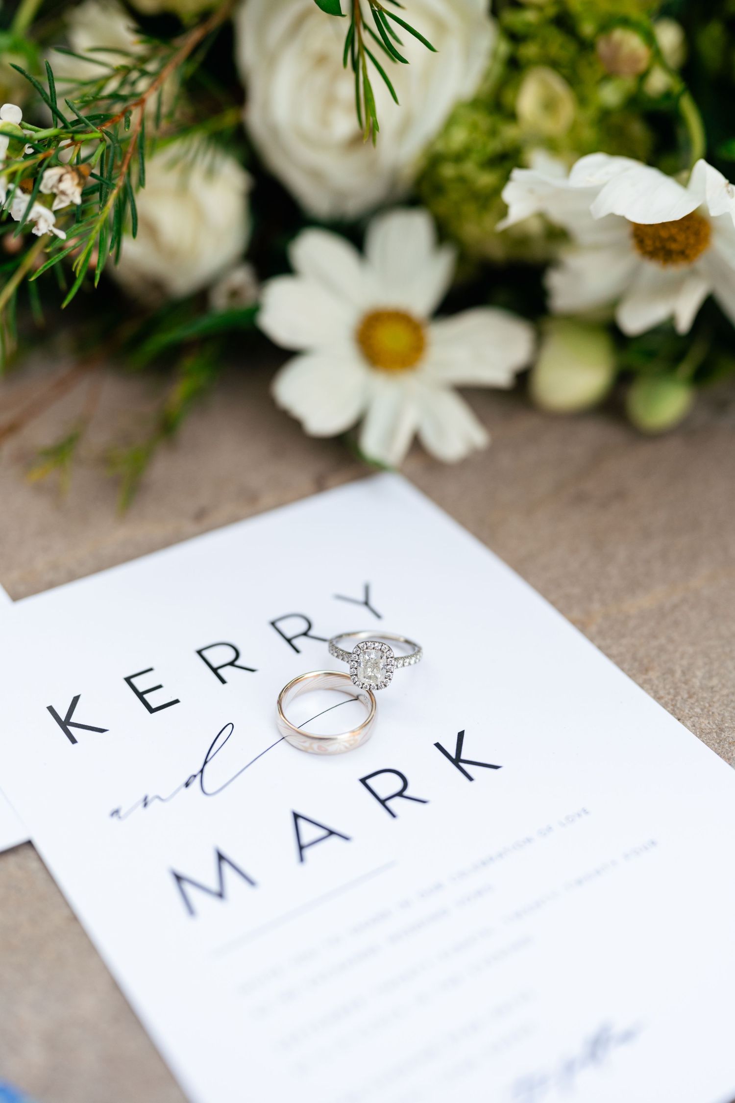 Wedding place cards with names and engagement ring displayed among white daisies and greenery.