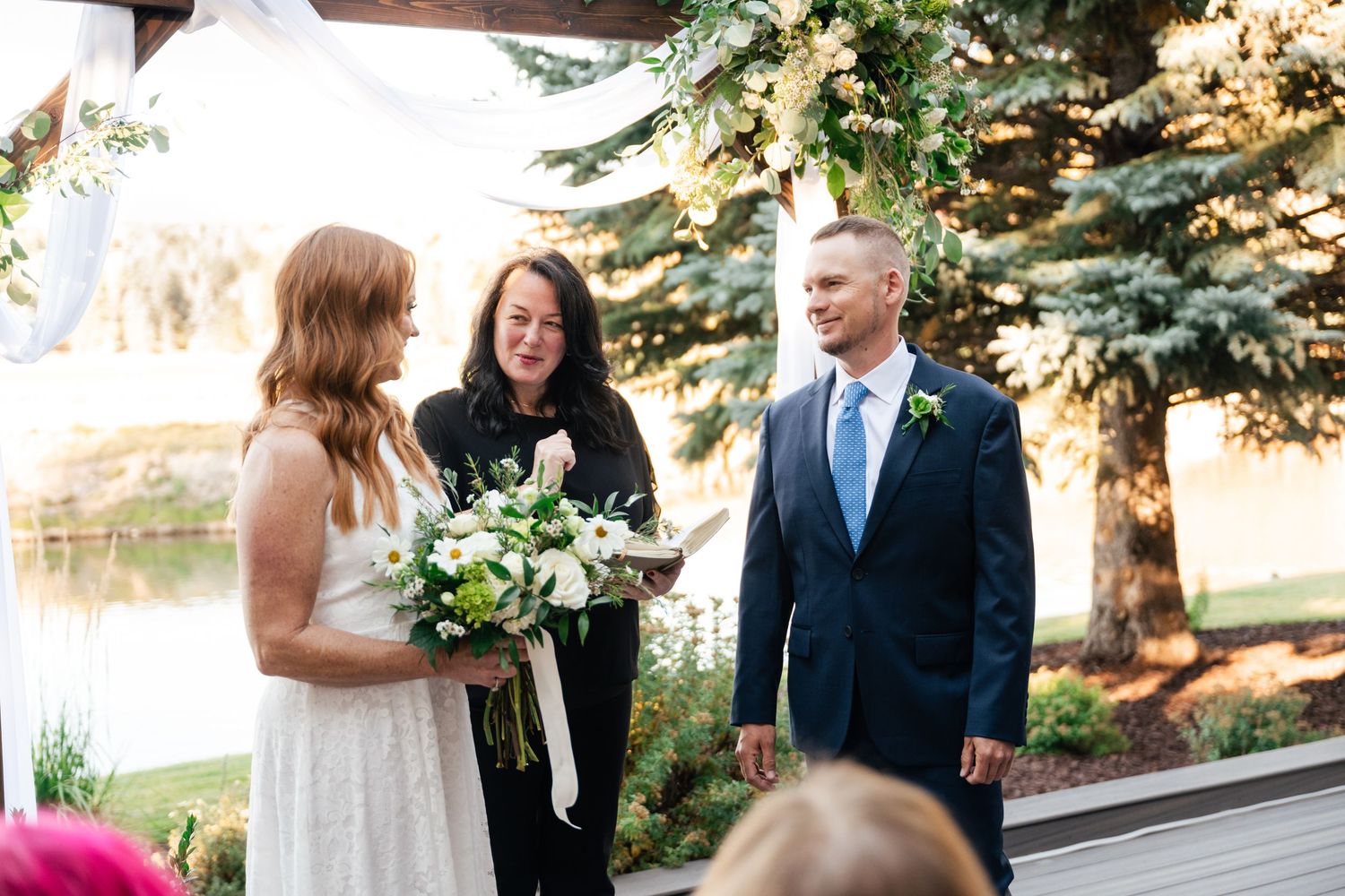 Outdoor wedding ceremony takes place on wooden deck overlooking serene lake with evergreen trees in background.