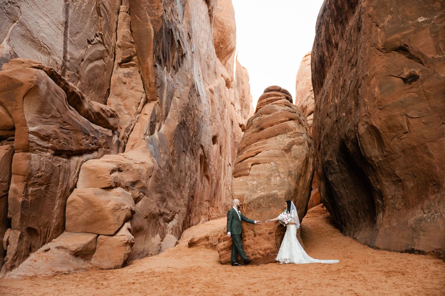 A couple in wedding attire stands between towering red sandstone walls in a dramatic desert canyon landscape.