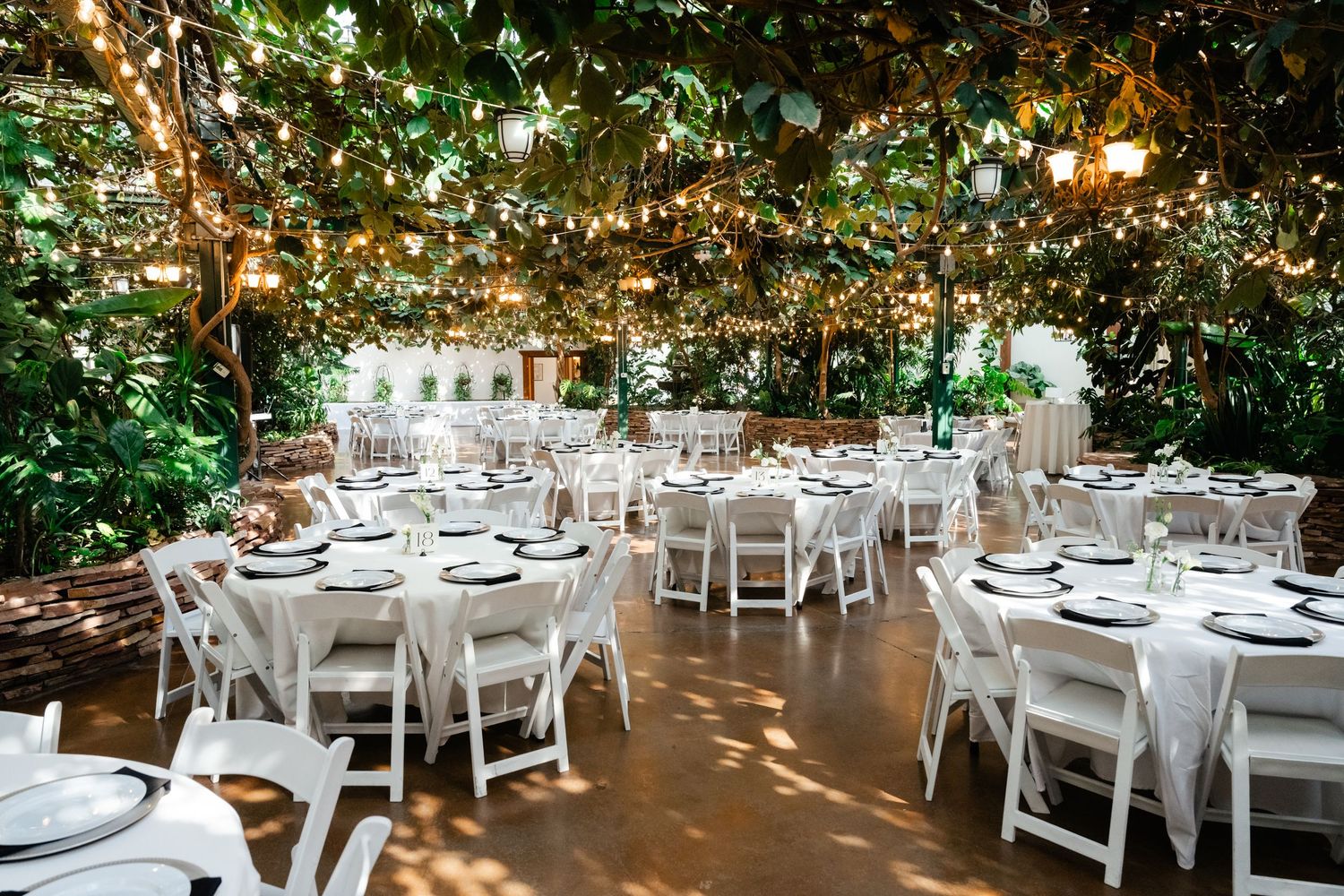 Elegant garden wedding reception setup with white tables and chairs under a canopy of twinkling string lights. Highland Gardens.