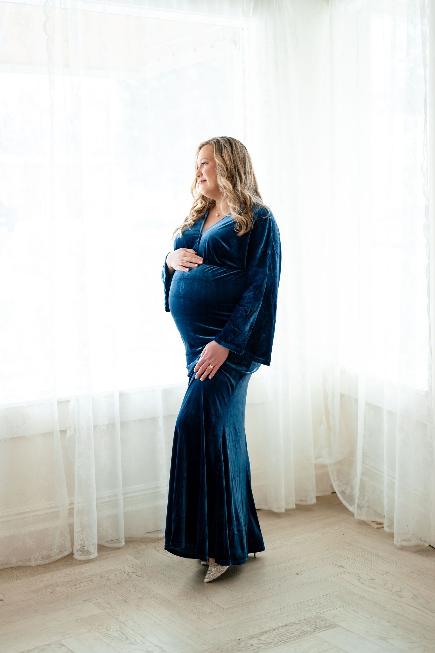 Someone wearing a navy blue dress poses gracefully near sheer white curtains in a bright minimalist room.