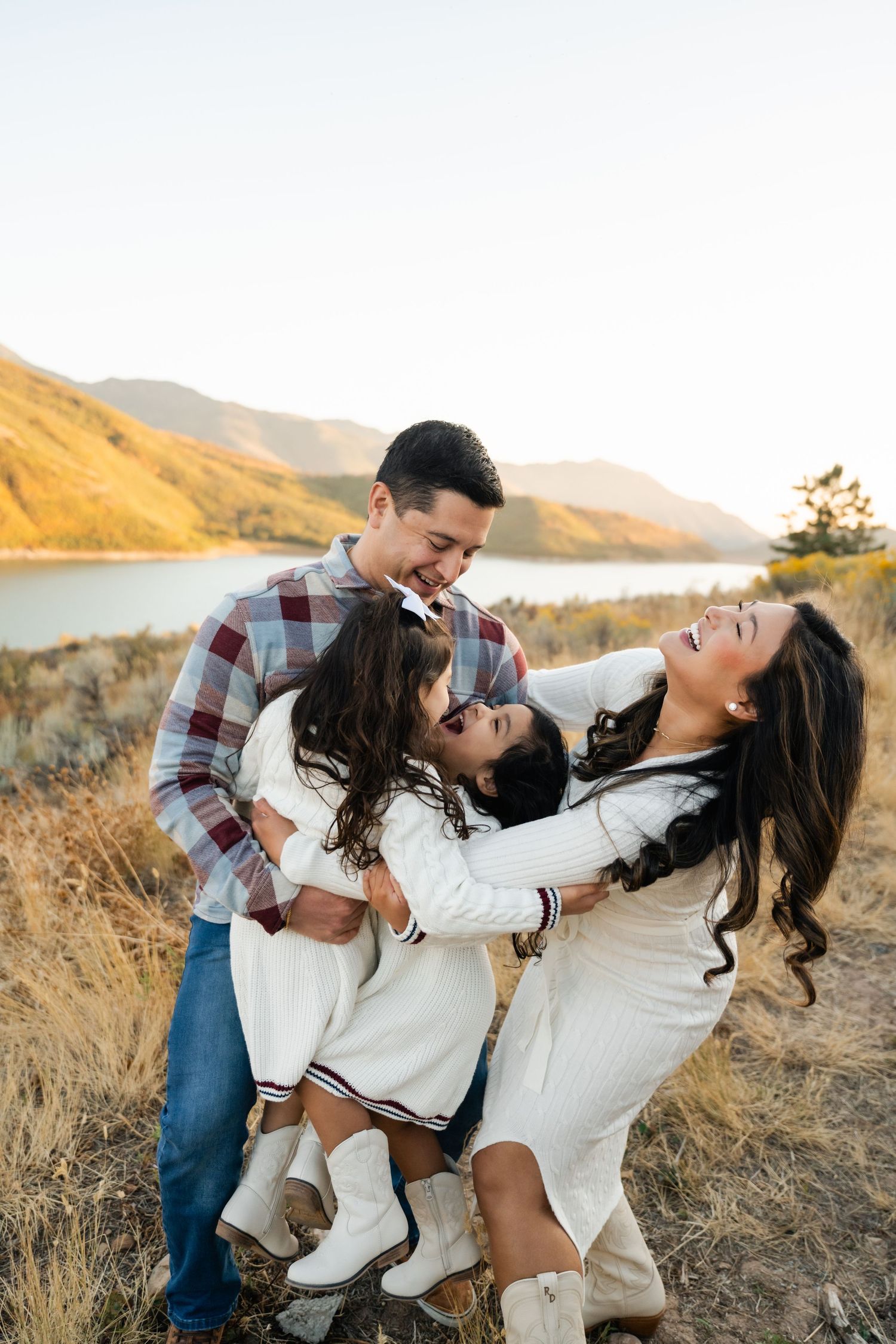 Romantic couple in casual attire embraces and laughs together during golden hour photoshoot by a scenic mountain lake.