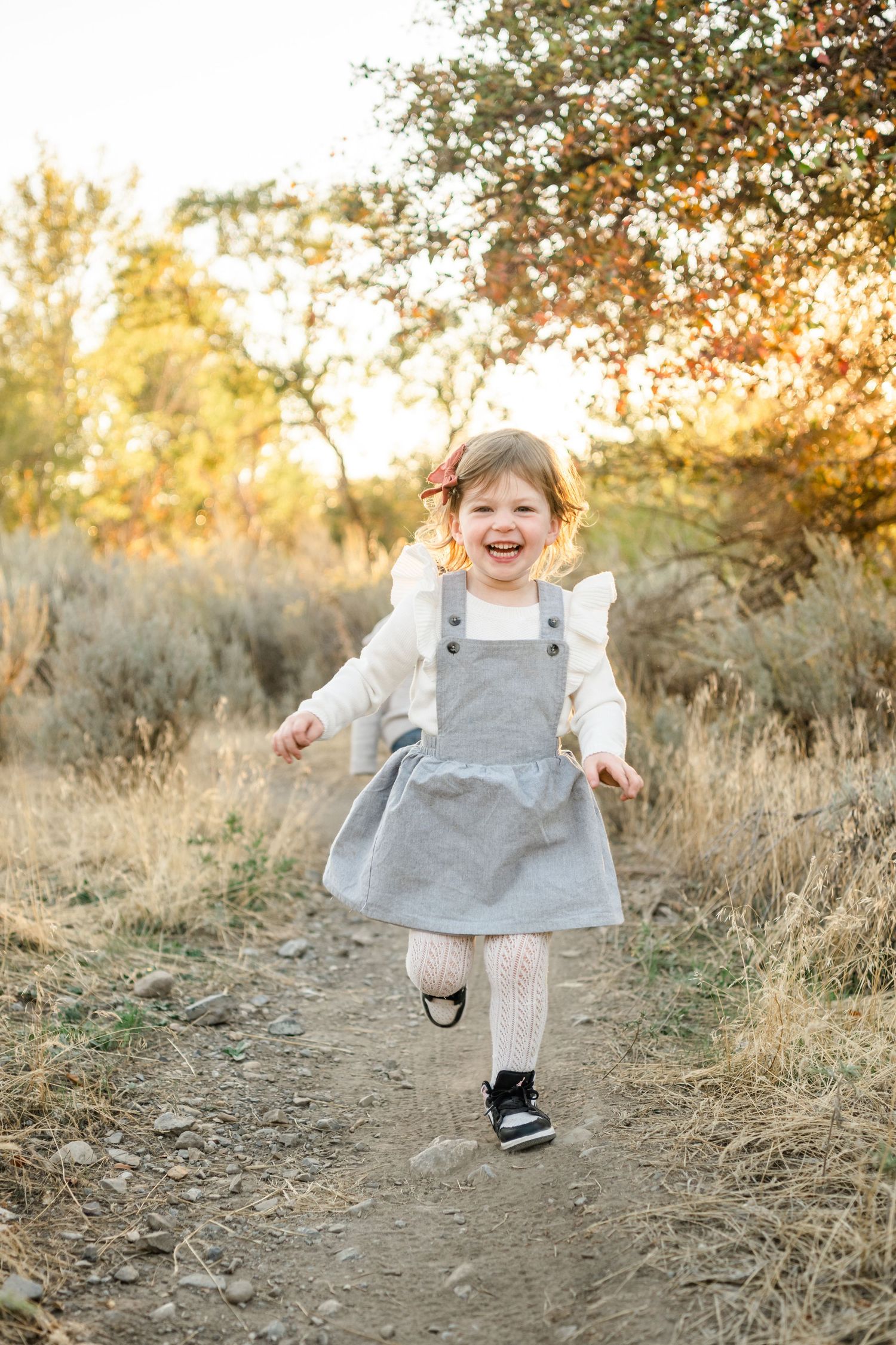 A child in a gray pinafore dress and white tights skips joyfully down a dirt path on an autumn afternoon.
