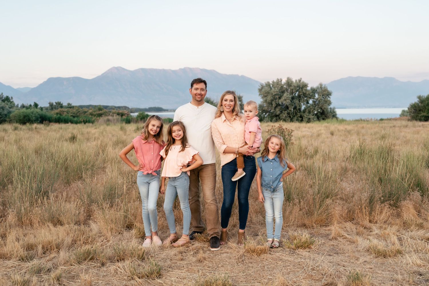 Family wearing coordinated coral and blue outfits poses together in a rustic field with mountains in background at sunset.