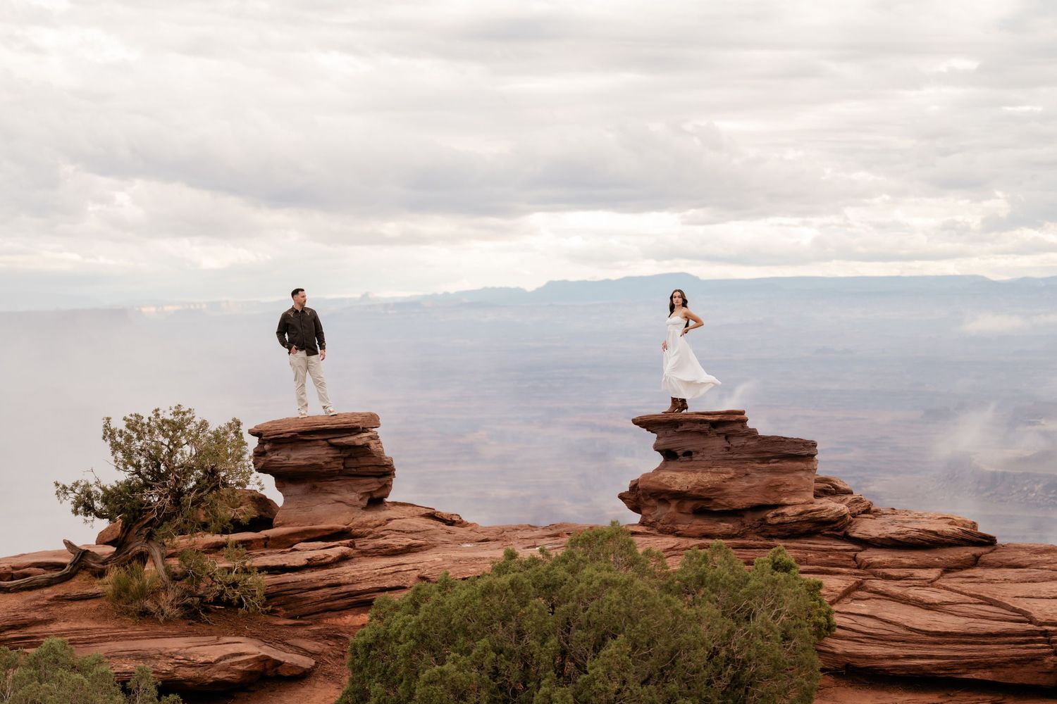 People stand on dramatic red rock formations overlooking a vast desert landscape at sunset.