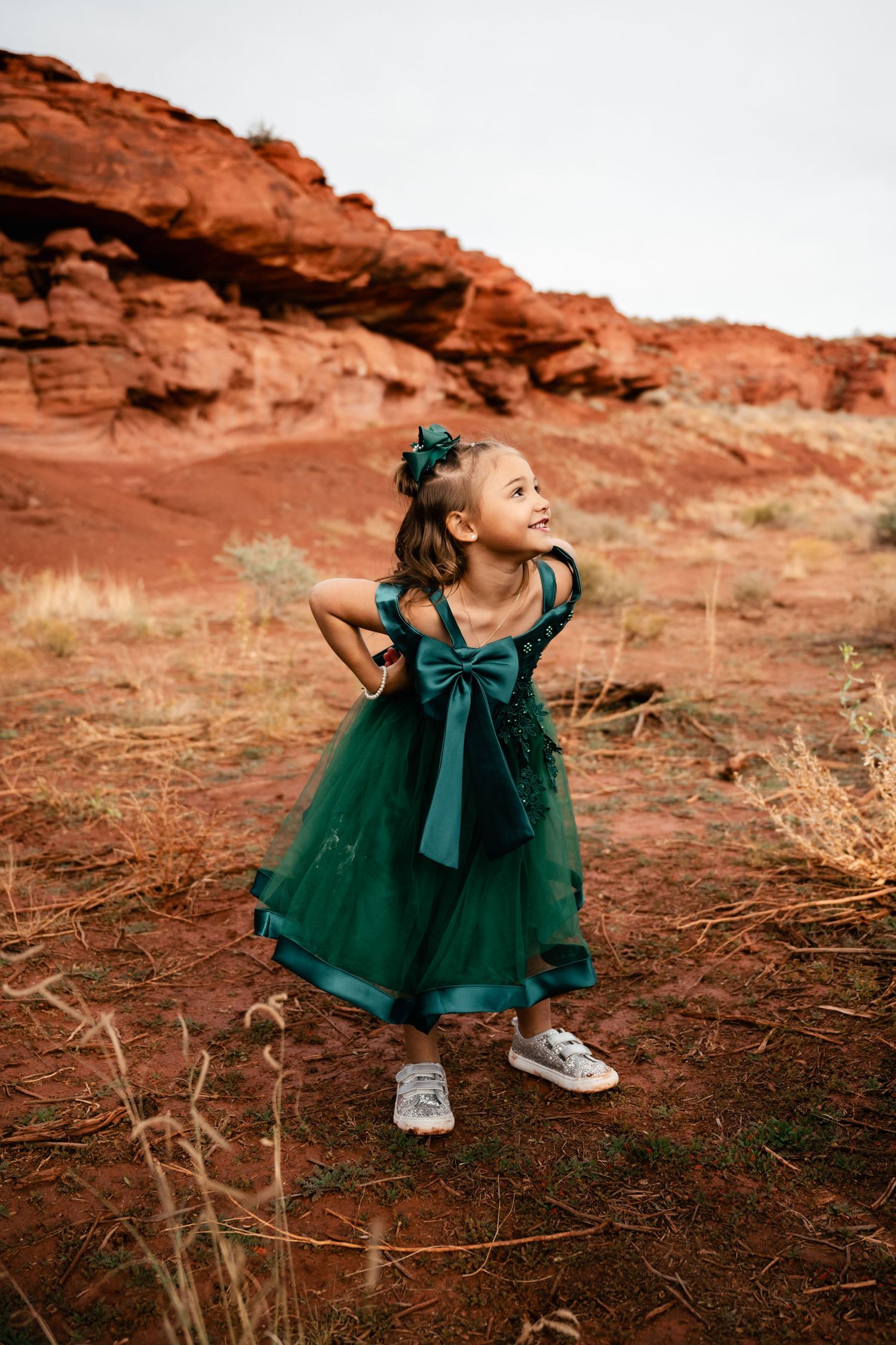 A small figure in an emerald green dress twirls joyfully against a backdrop of red rock formations in a desert landscape.