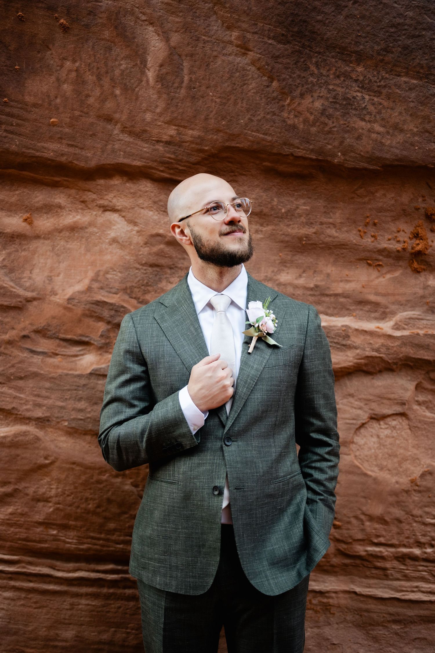 A person in a gray suit adjusts their tie while standing against a red rock wall backdrop.