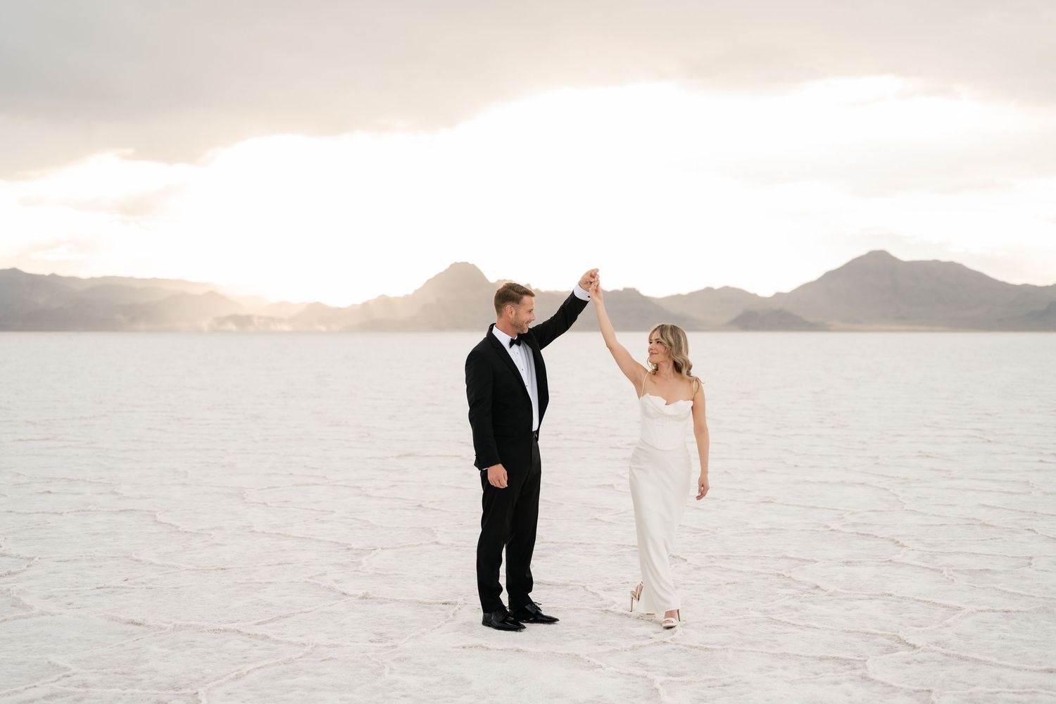 Salt Flats Elopement Photographer.A couple in wedding attire dance together on a white salt flat with mountains in the background at sunset.