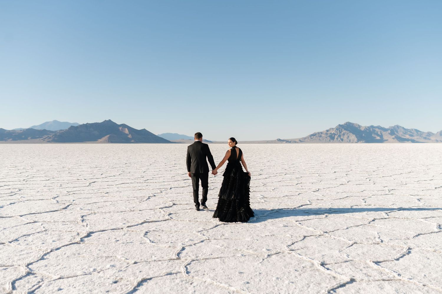 Silhouettes of people sitting on vast white salt flats with mountains in the distance under a bright blue sky. Salt Flats Photographer.