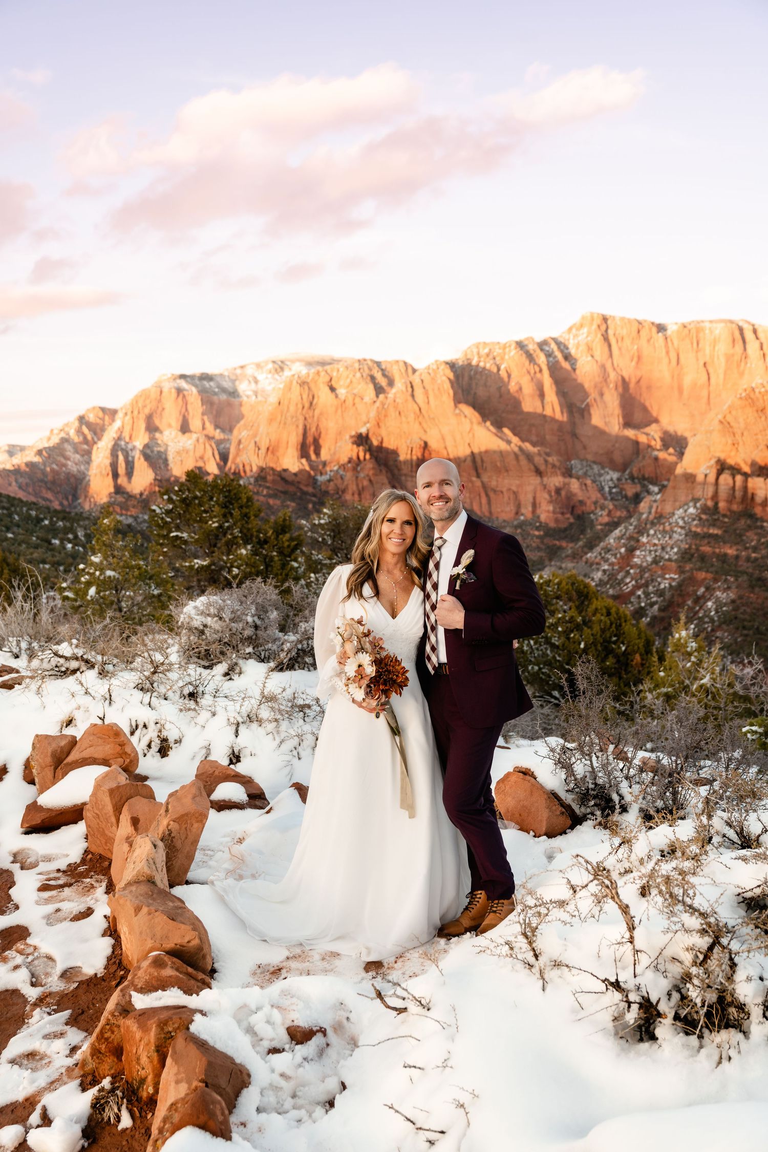 Newlyweds pose in a snowy desert landscape with towering orange cliffs and winter vegetation.