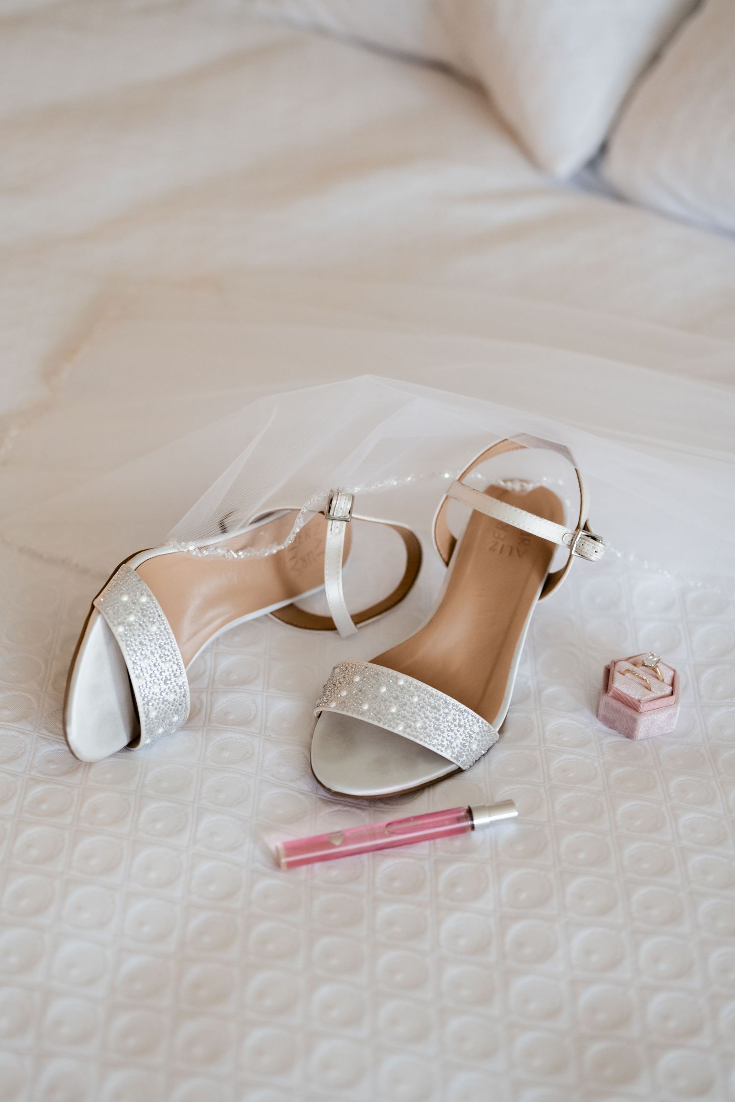White bridal sandals with rhinestone details and lip gloss lay on a textured white bedspread.