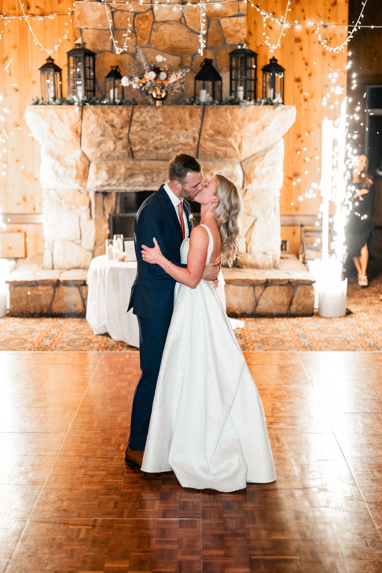 Romantic first dance moment under string lights with a stone fireplace backdrop.