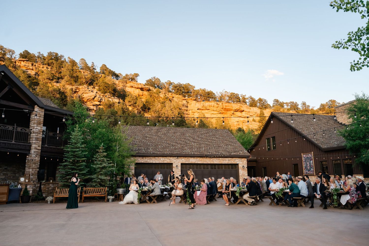 Panoramic view of a rustic lodge venue nestled against golden cliffs at sunset with guests gathered outside.