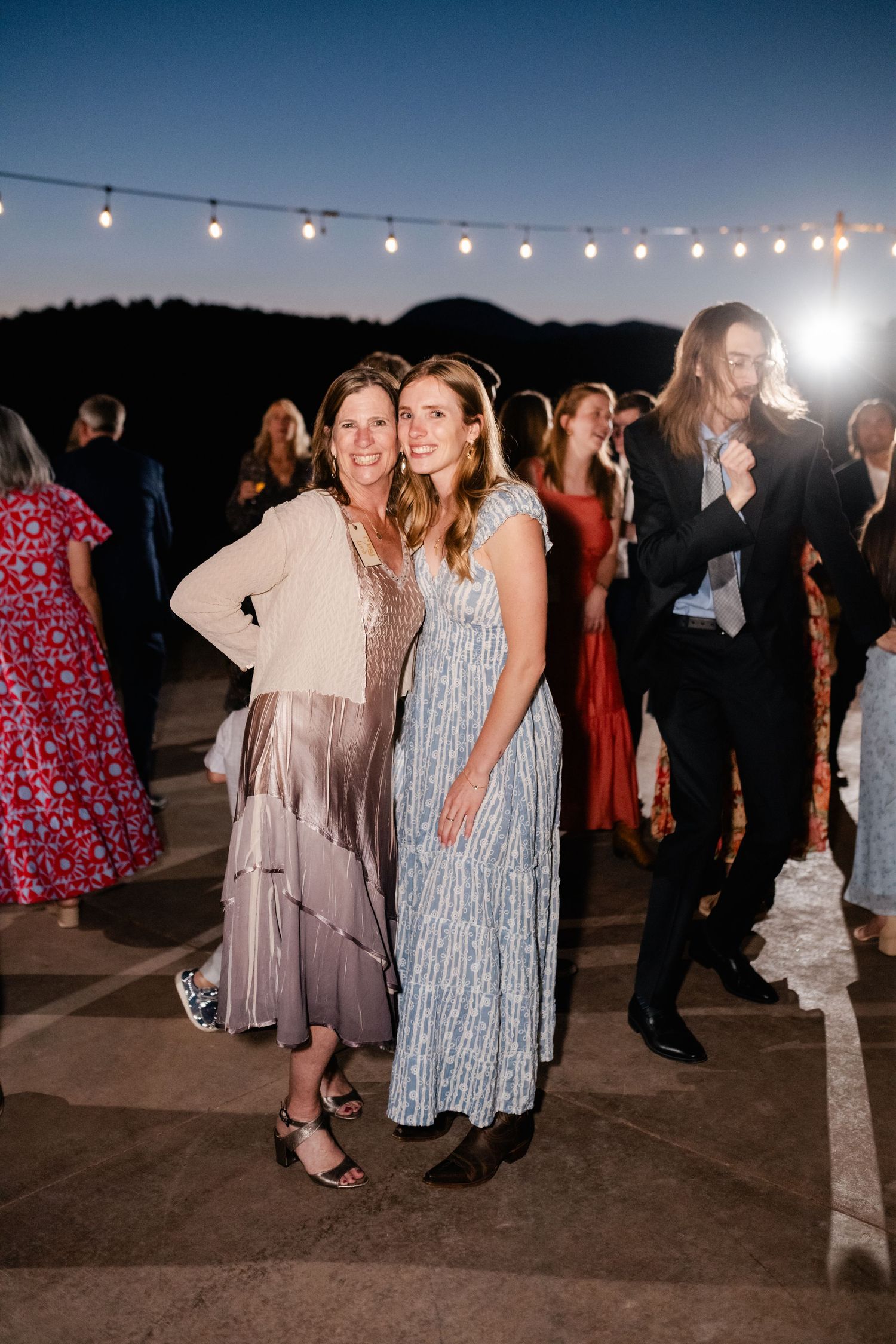 Wedding guests in colorful dresses gather on outdoor patio under string lights at dusk.