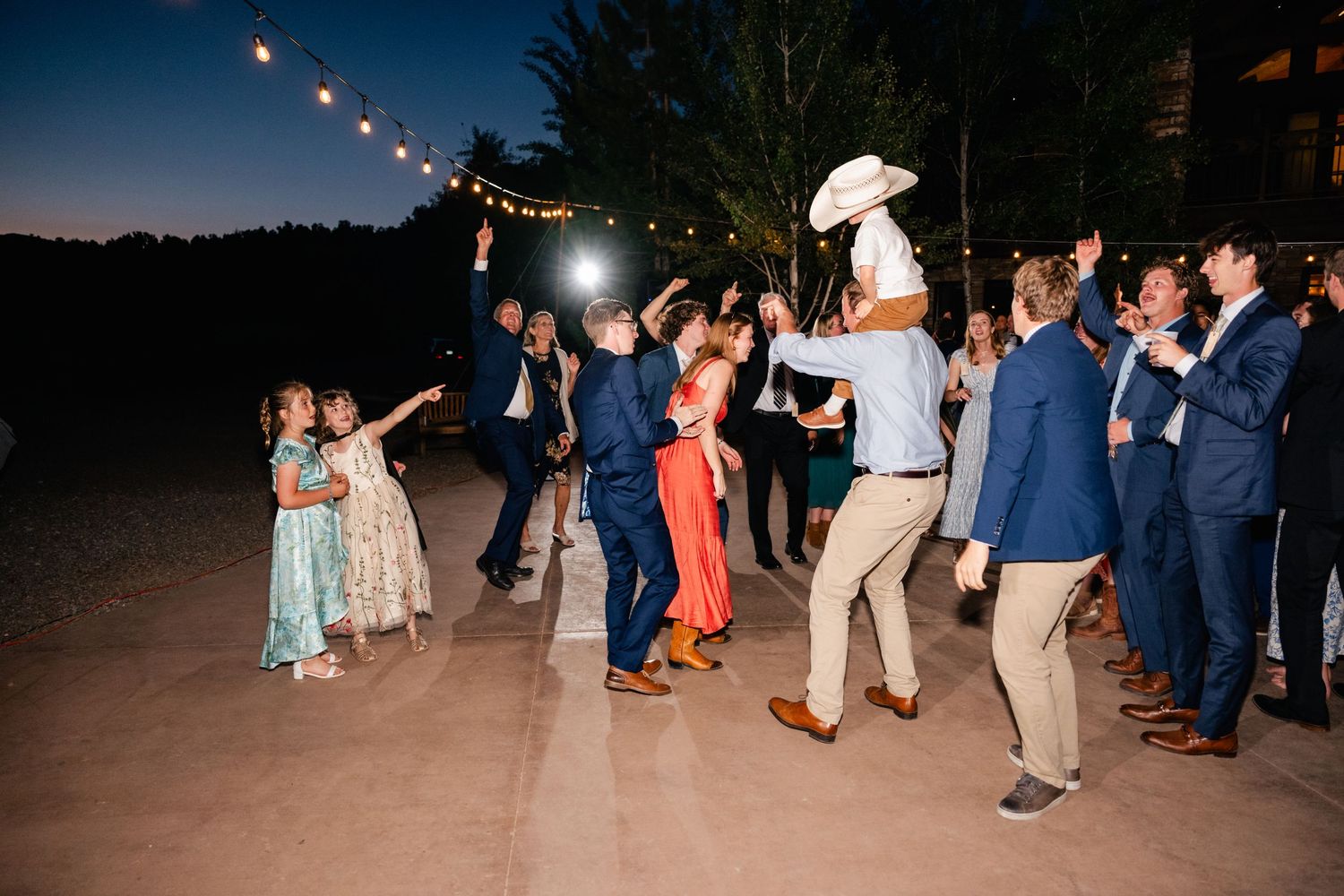 String lights illuminate an outdoor dance floor filled with celebrating wedding guests at dusk.