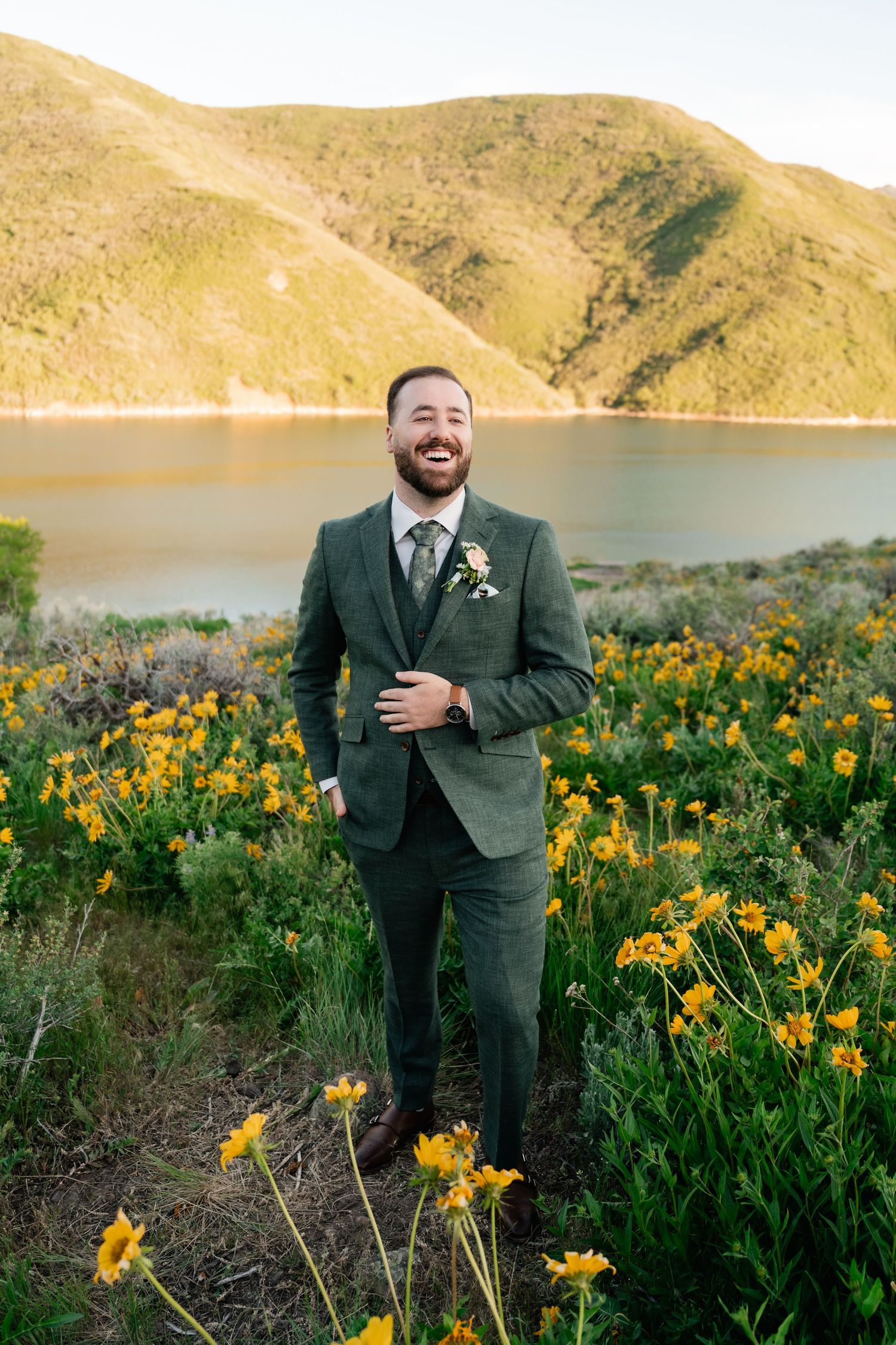 A groom in dark green suit poses among yellow wildflowers with a lake and mountains behind him.