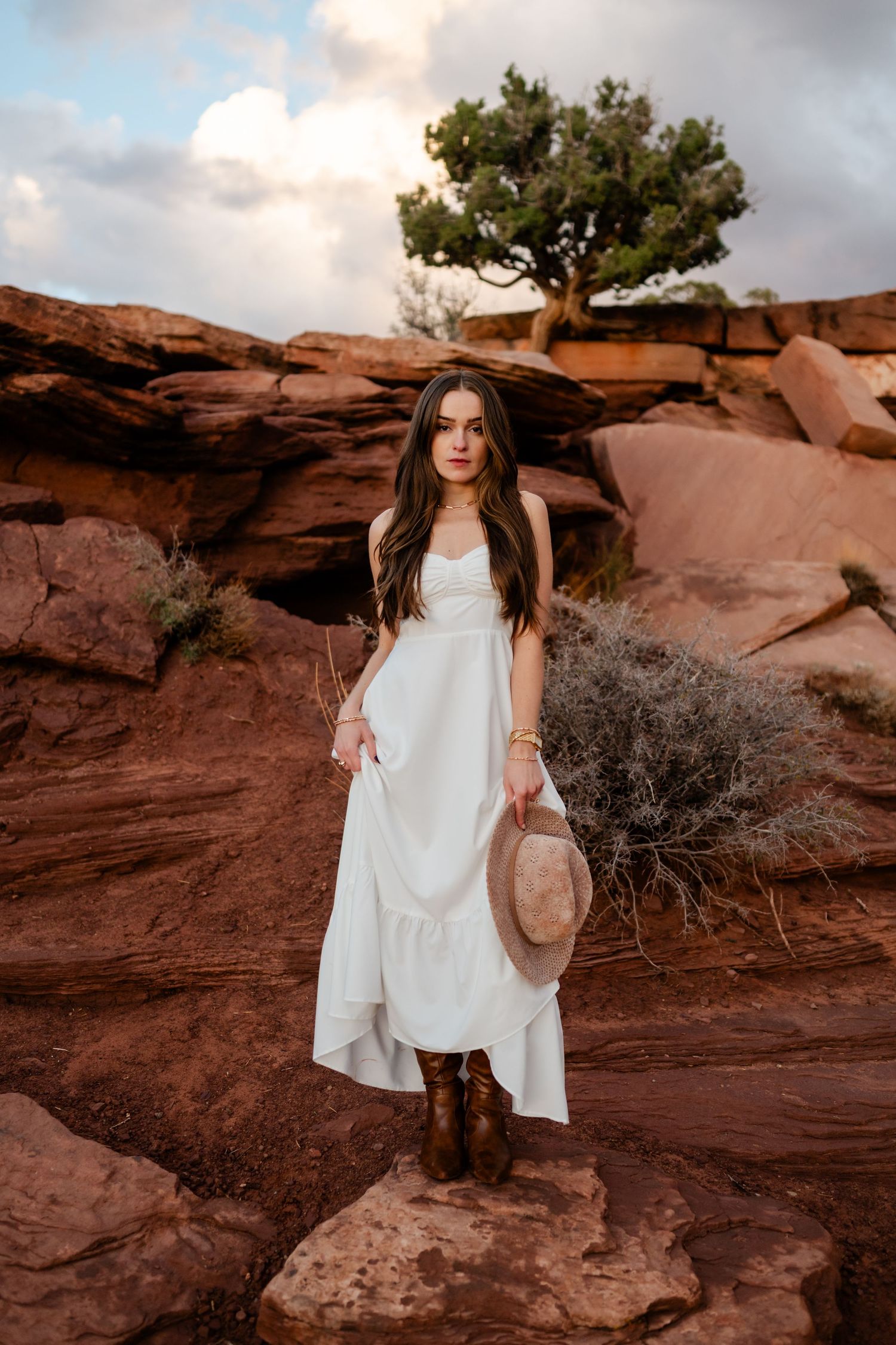 Woman in flowing white dress and boots standing on red rock formations in a desert landscape.