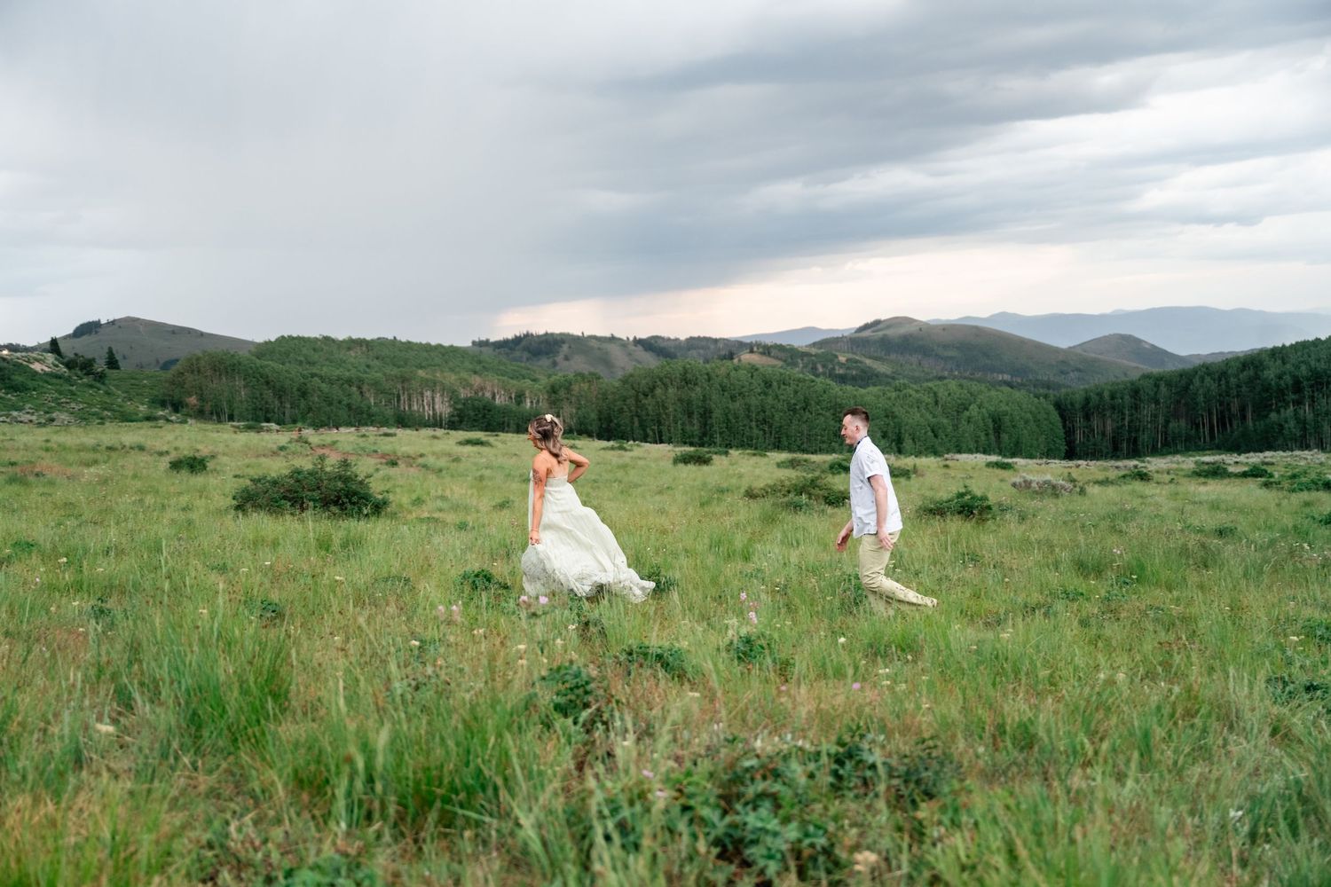 Two people in white outfits walking through a mountain meadow under dramatic cloudy skies.