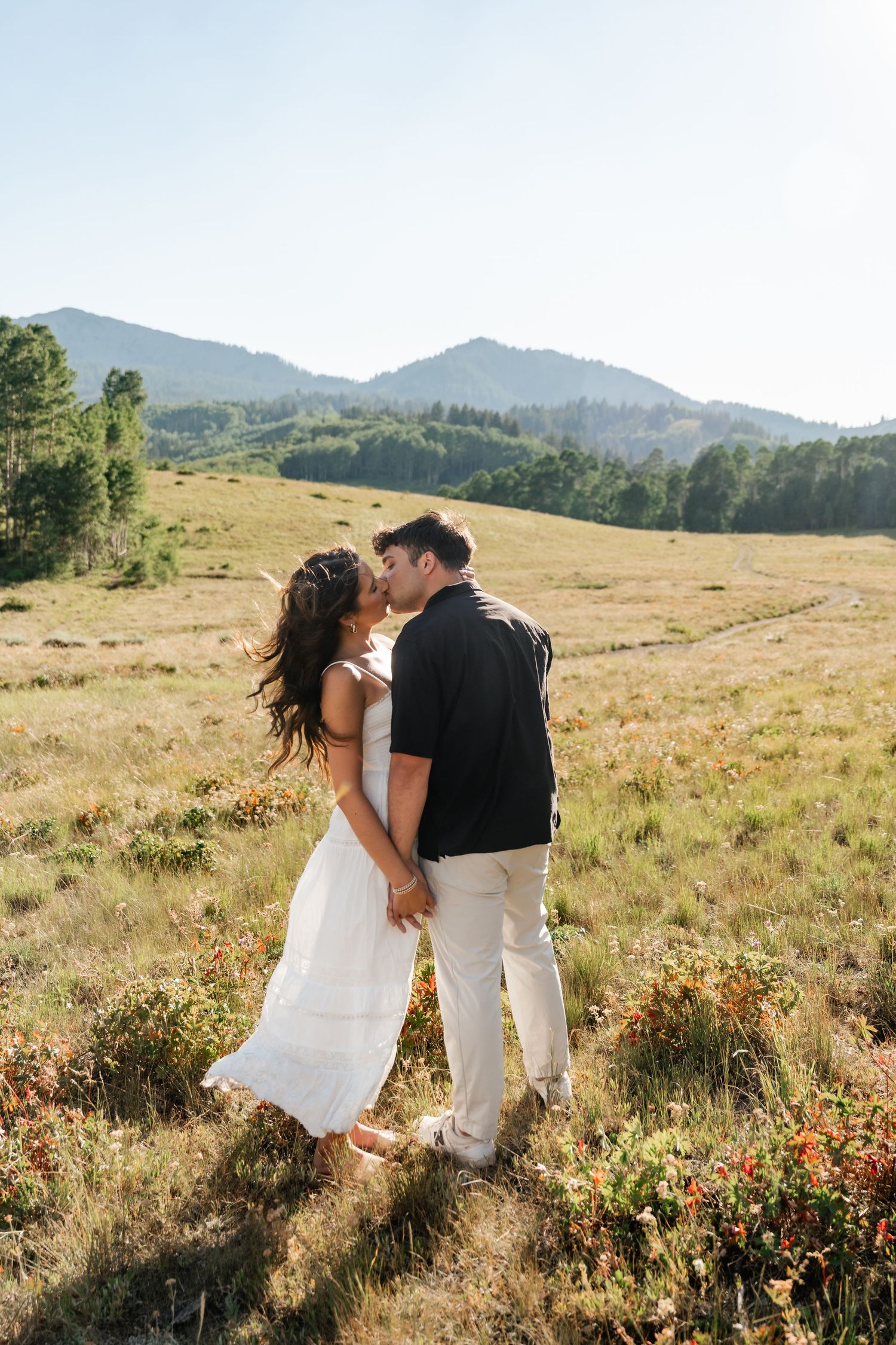 Couple shares a romantic moment in a golden mountain meadow with wildflowers during a sunny proposal photoshoot. Utah Proposal Photographer.