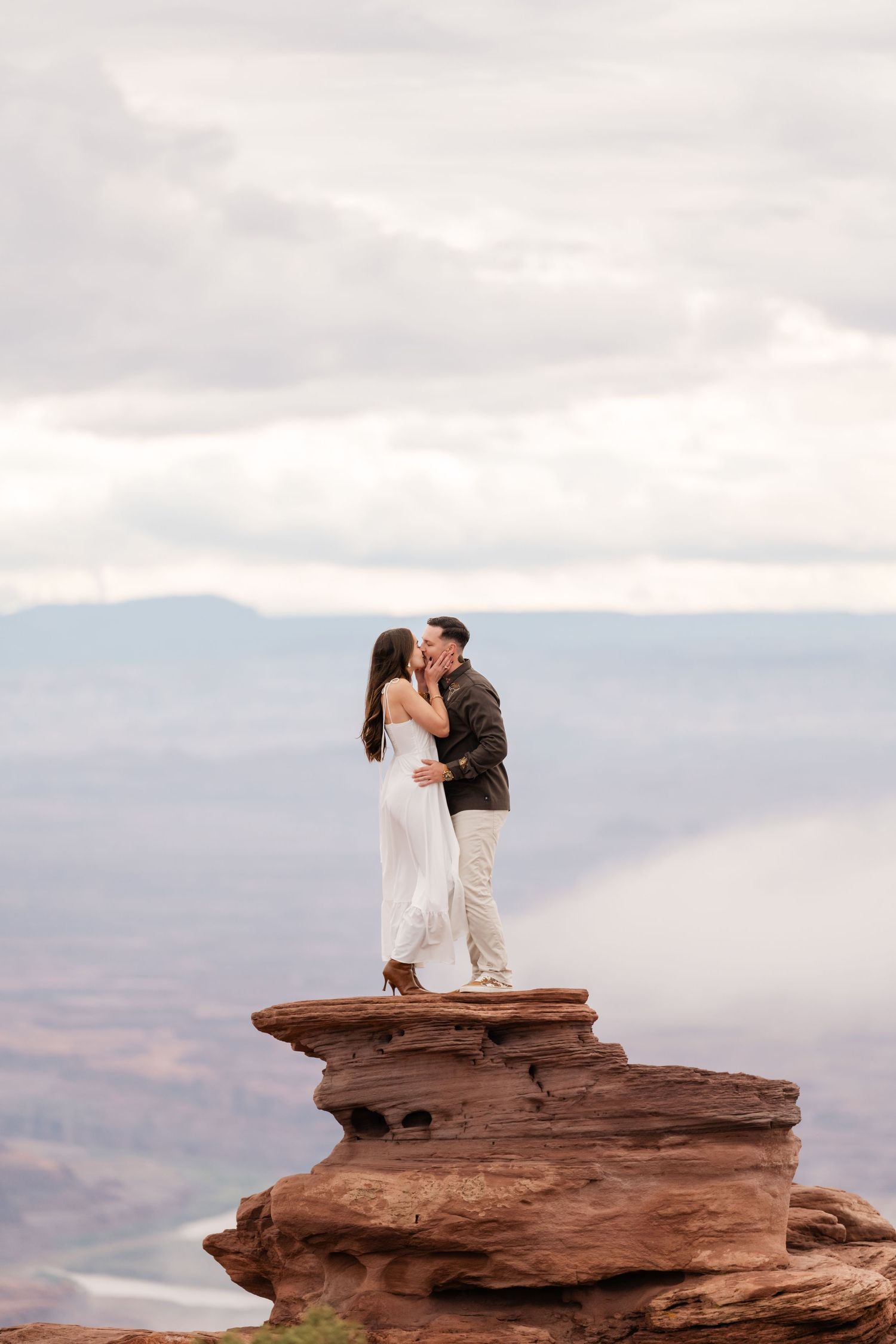 A couple shares a romantic kiss while standing on a dramatic red rock formation overlooking a desert landscape at sunset in Moab, Utah.