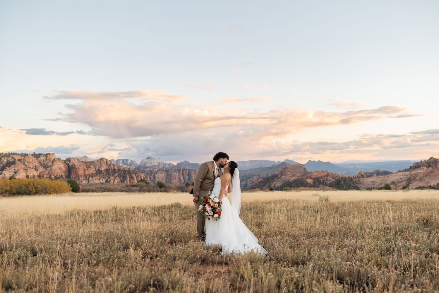A panoramic view of a wedding photoshoot in an open field with mountains and sunset clouds in the distance.