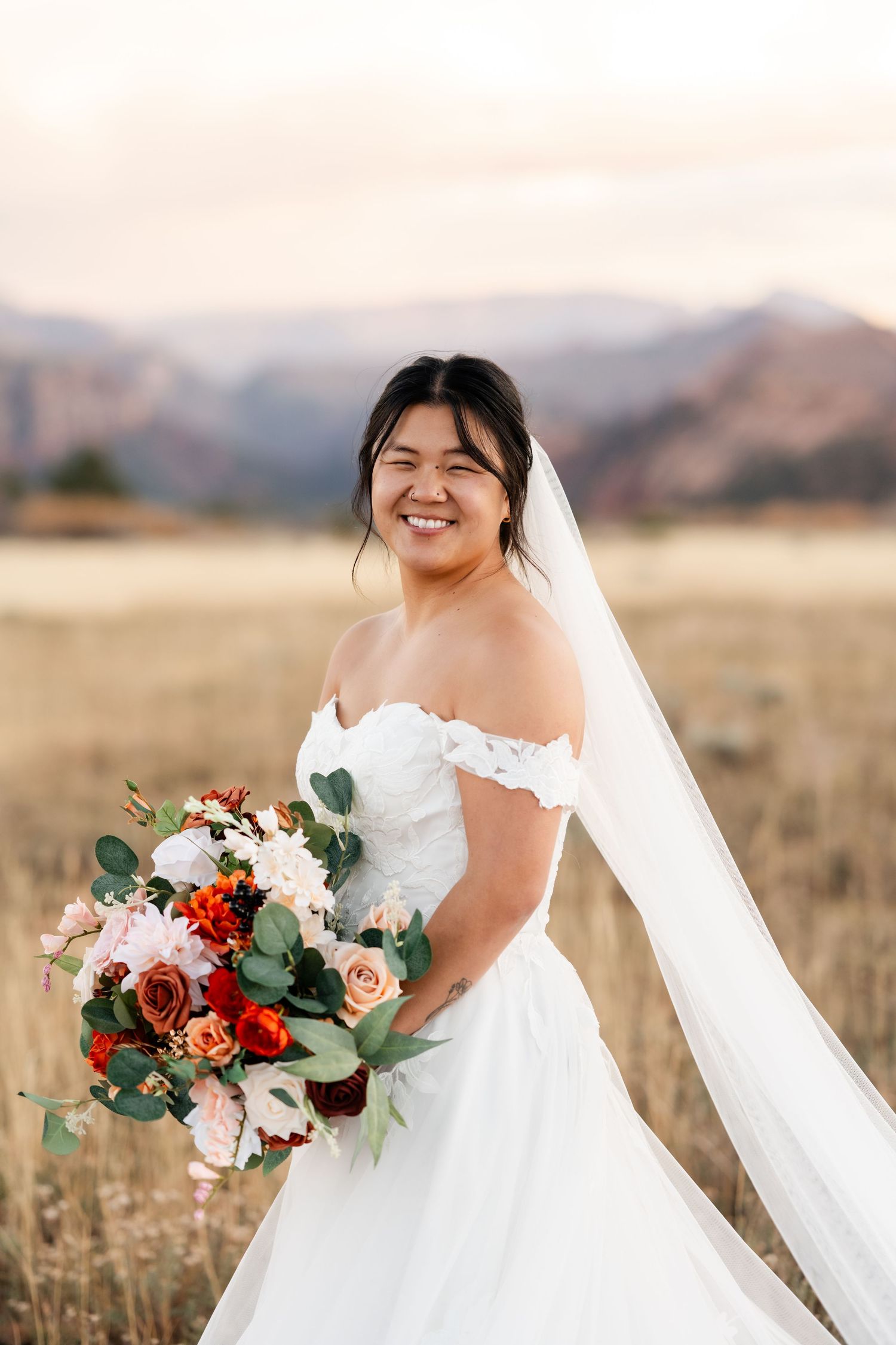 A bride in a white gown holds a vibrant bouquet of pink and red flowers with eucalyptus in an outdoor setting.