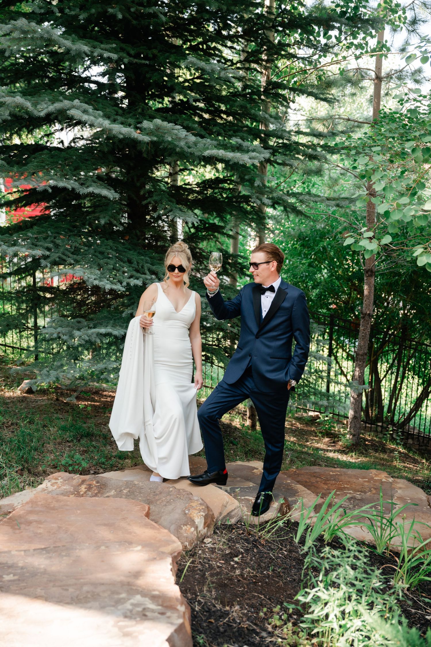 Wedding couple poses playfully on rocky landscaping surrounded by evergreen trees and natural scenery.
