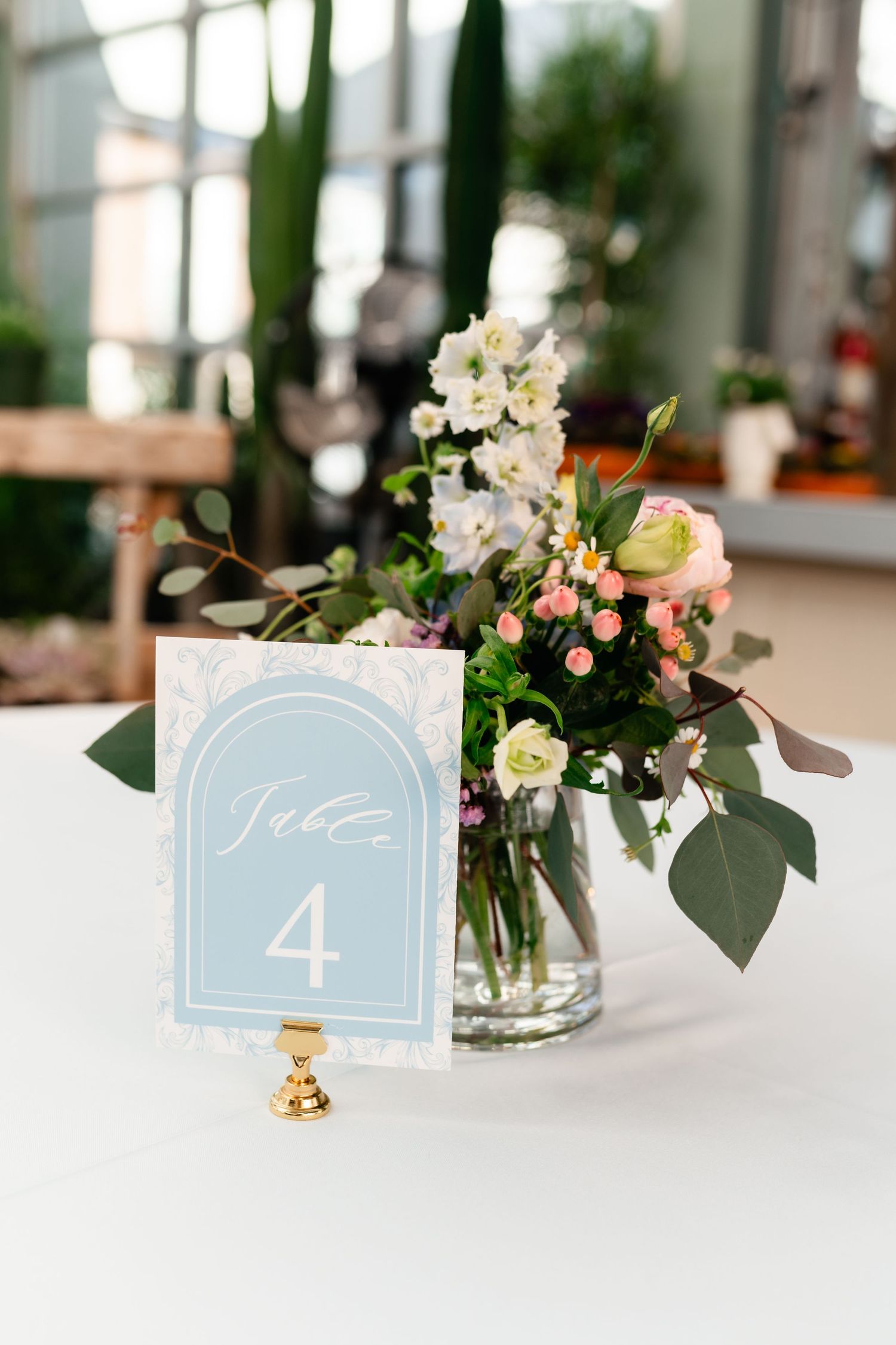Light blue wedding table number card with floral centerpiece in glass vase on white tablecloth.