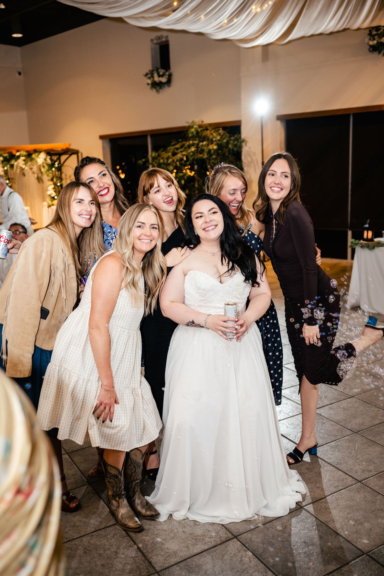 A joyful group of friends pose together at a wedding reception with white decor and twinkling lights in the background.