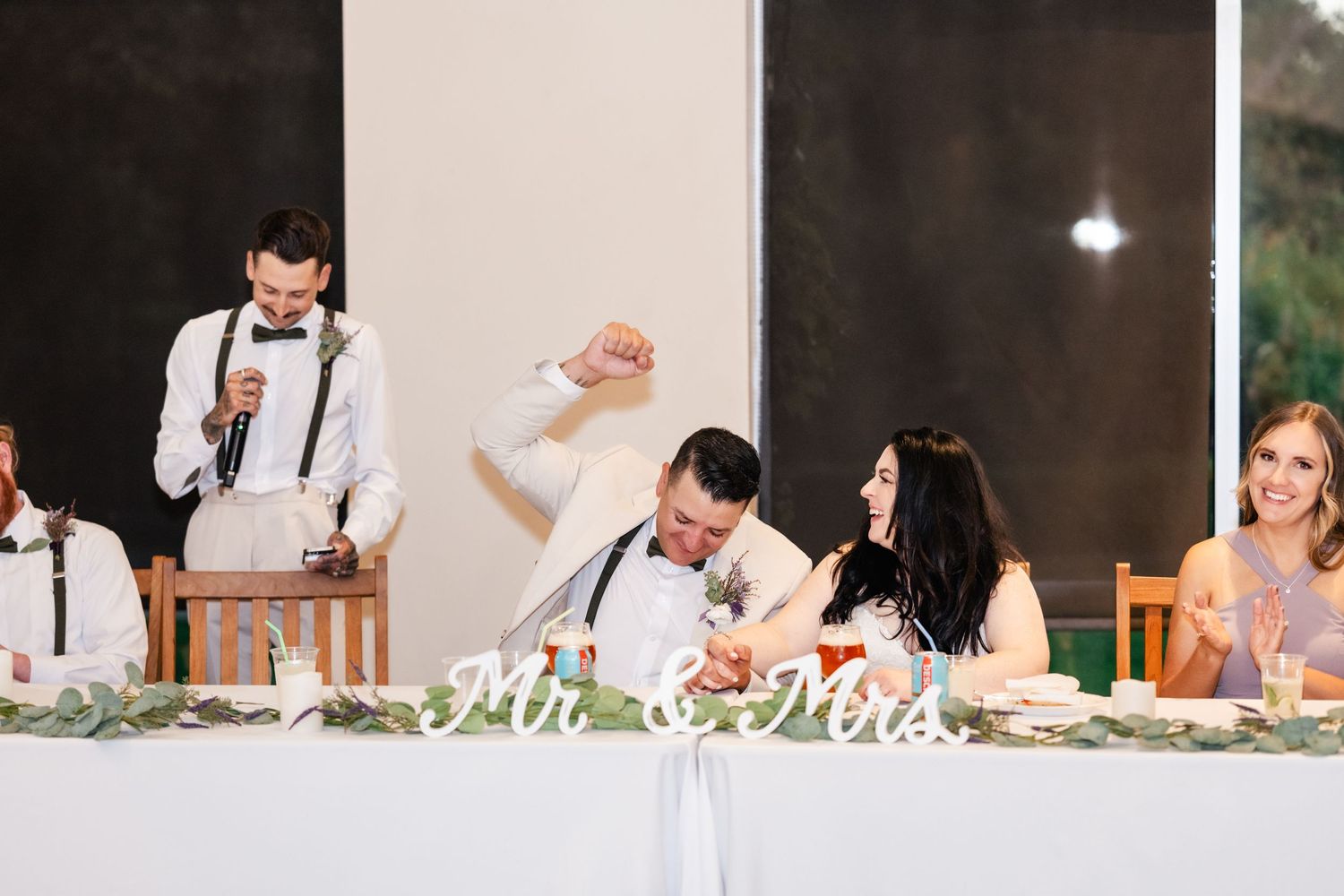 Wedding guests at a reception head table laugh and celebrate during dinner.