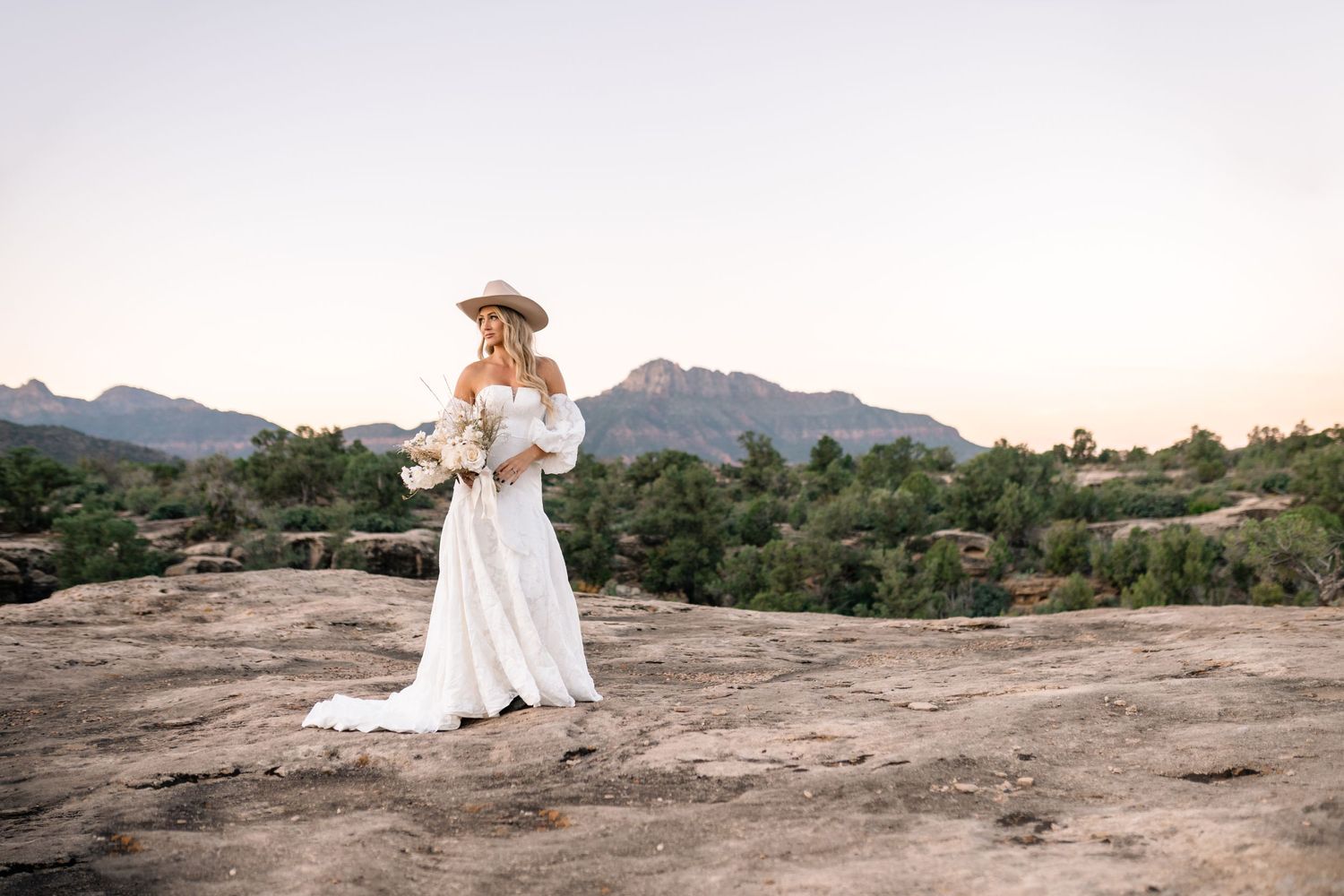 Bridal portrait in off-shoulder dress and cowboy hat against desert mountain scenery.