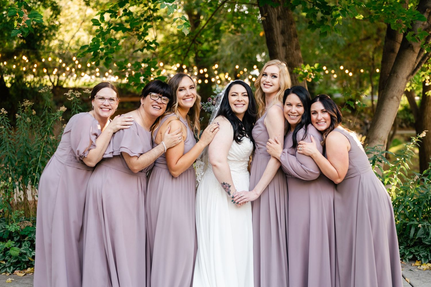 Wedding party wearing mauve dresses posing together outdoors under green trees.